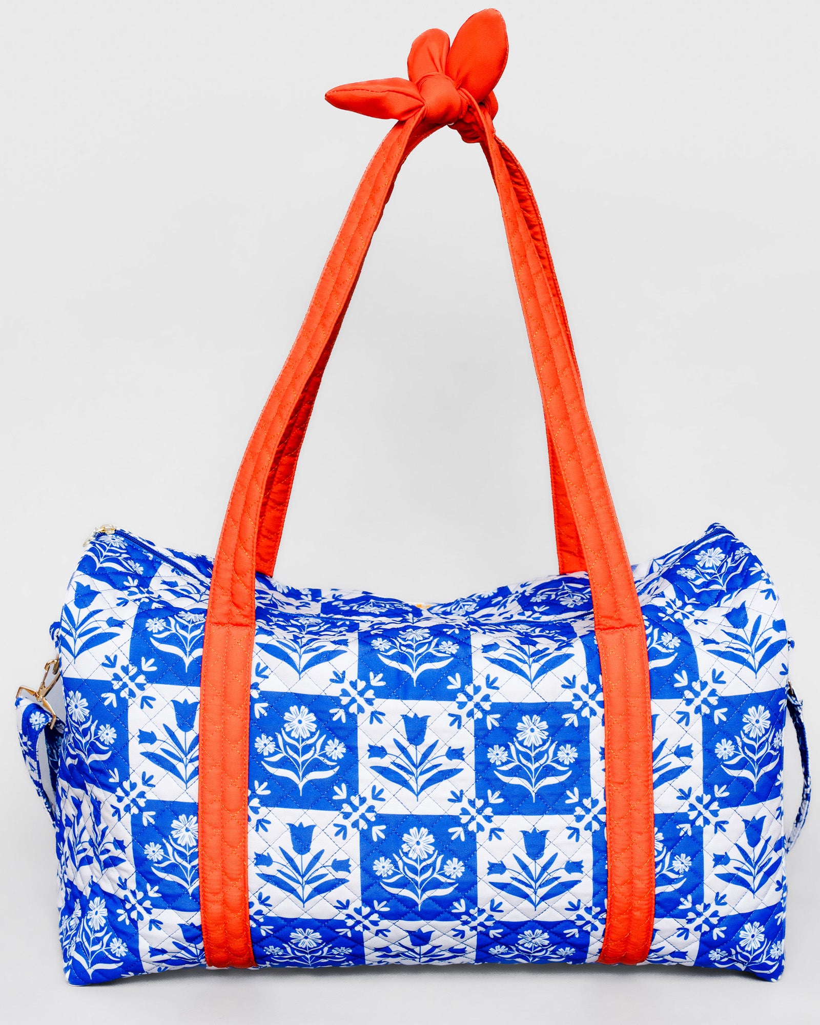 A duffle bag with a blue and white floral tile pattern and bright orange handles tied at the top, shown against a light gray background.