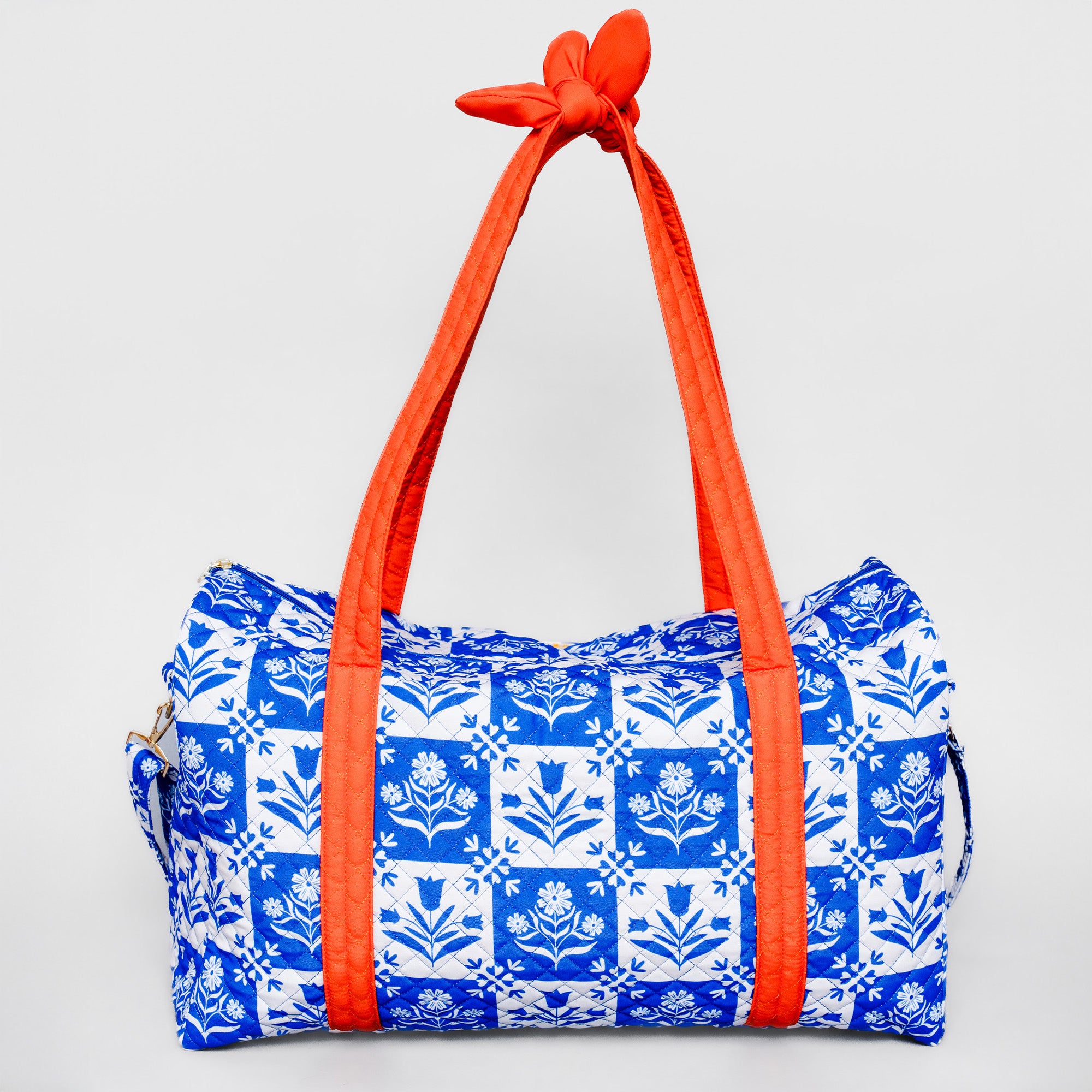 A duffle bag with a blue and white floral tile pattern and bright orange handles tied at the top, shown against a light gray background.