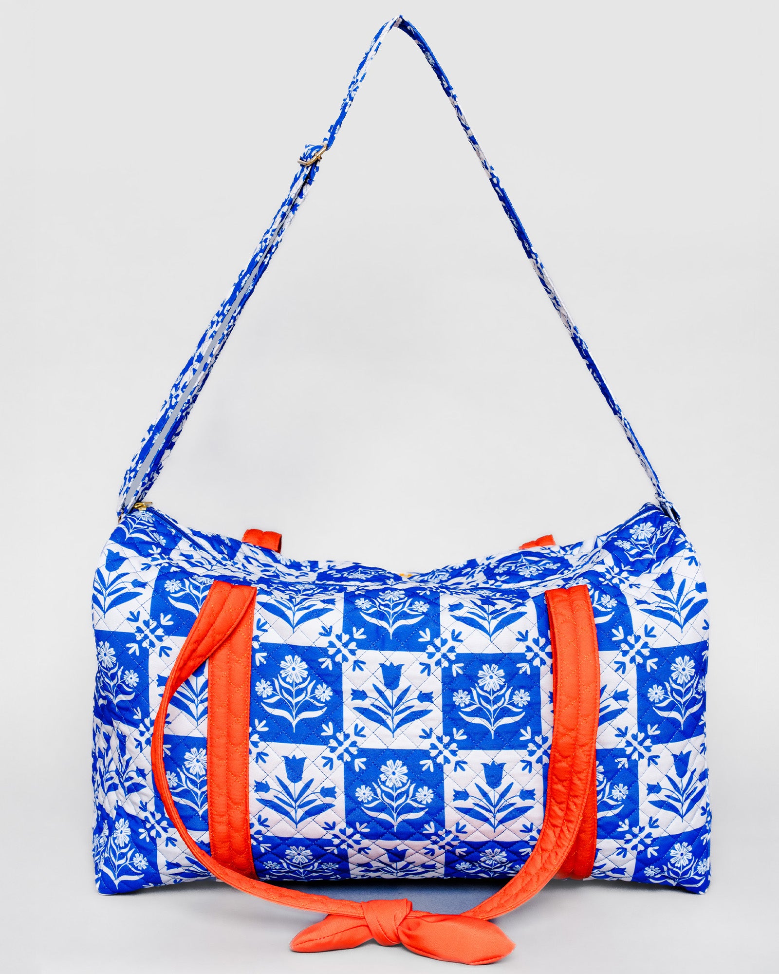 A blue and white patterned duffel bag with bright orange straps and a matching patterned shoulder strap, photographed against a light gray background.