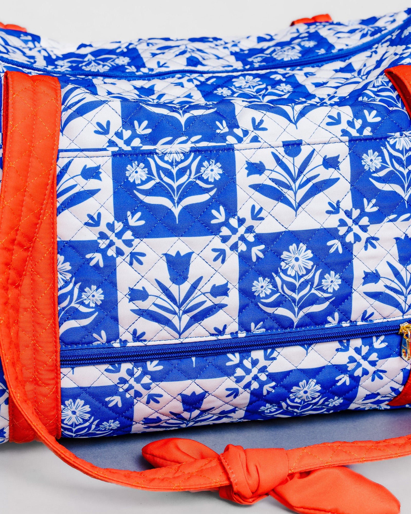 A quilted duffel bag featuring a blue and white floral block print pattern with bright orange straps and trim, shown against a light gray background.