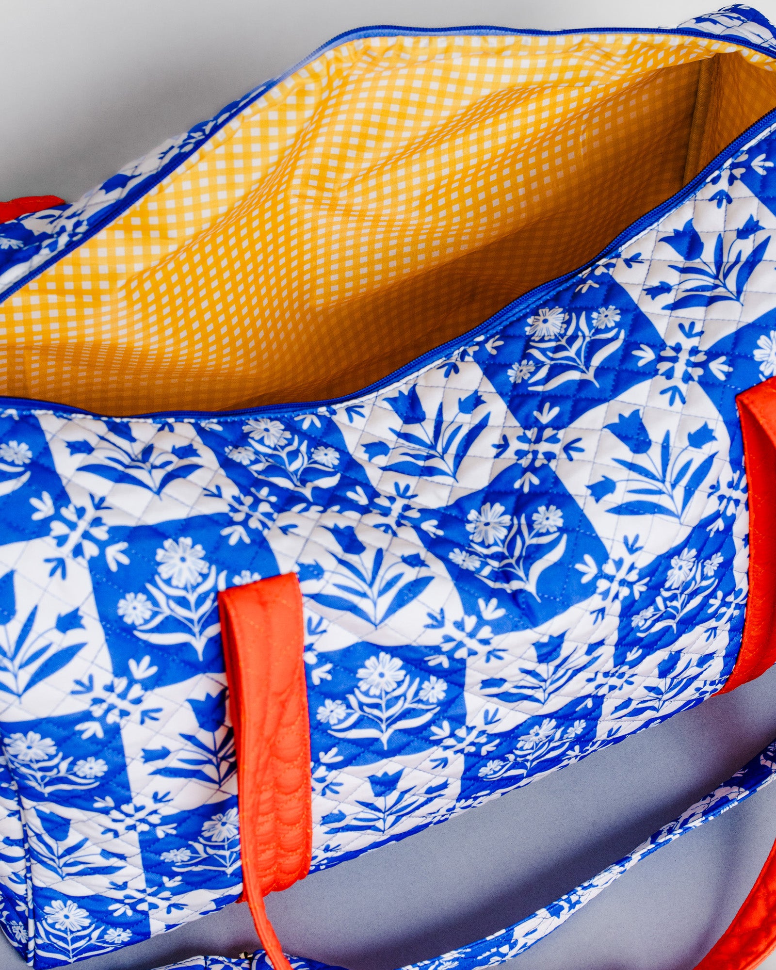 A quilted tote bag with a blue and white floral block-print pattern, a yellow gingham lining, and bright orange handles, shown on a neutral gray background.