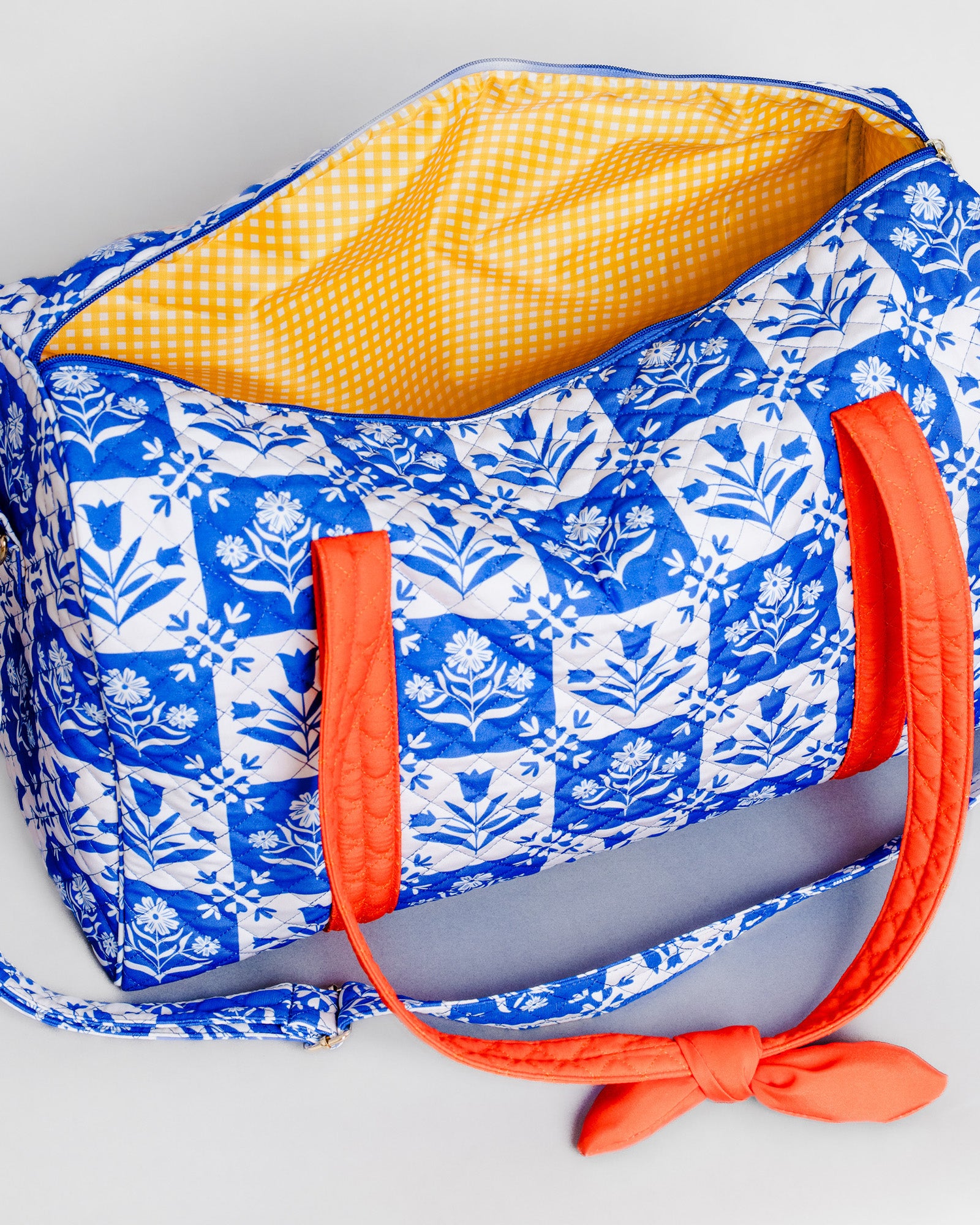 A blue and white patterned duffel bag with a bright yellow gingham interior and orange handles, shown on a light gray background.