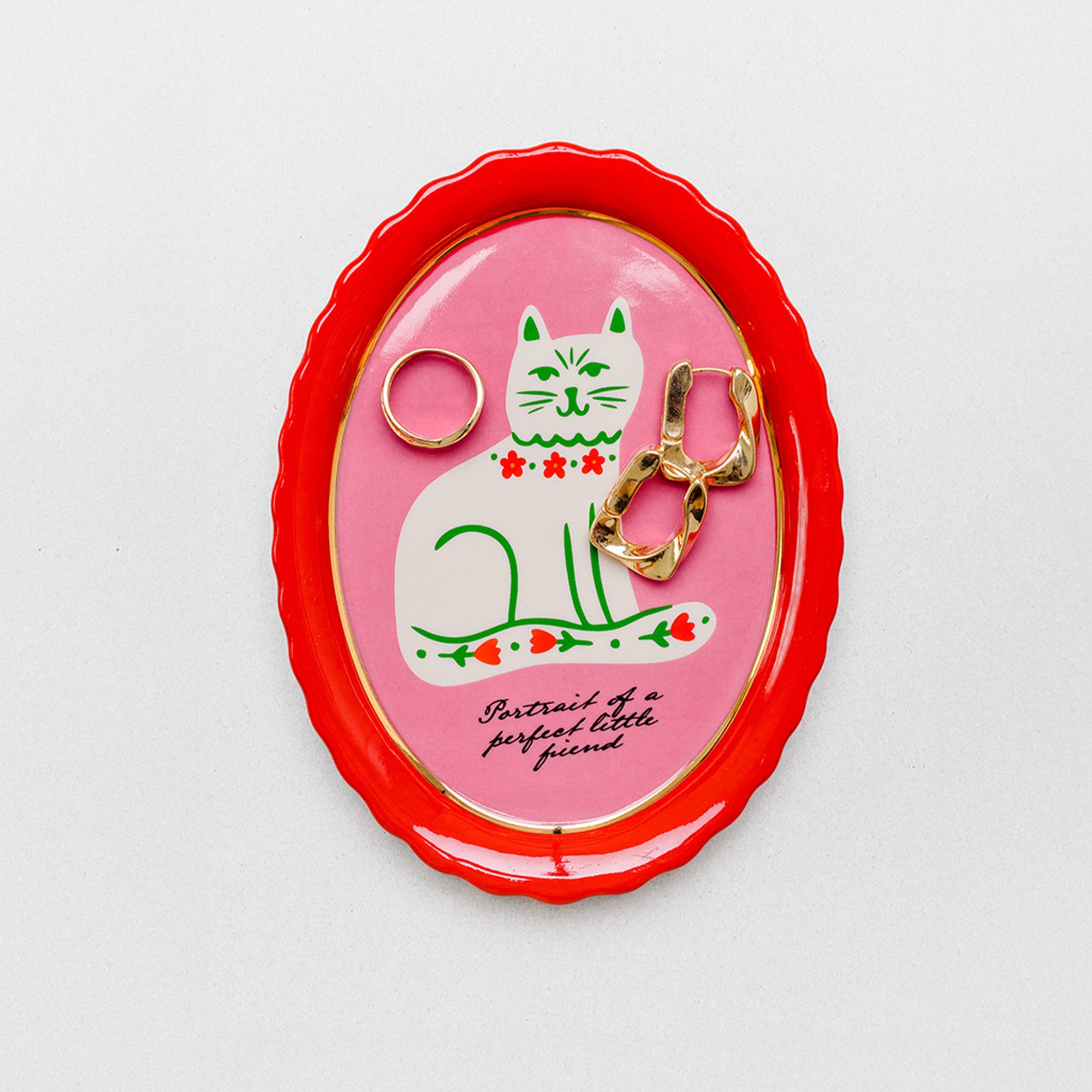 An oval enamel pin holder with a bright red scalloped border, featuring a pink center with a white cat illustration and gold-tone jewelry displayed inside, set against a clean white background.