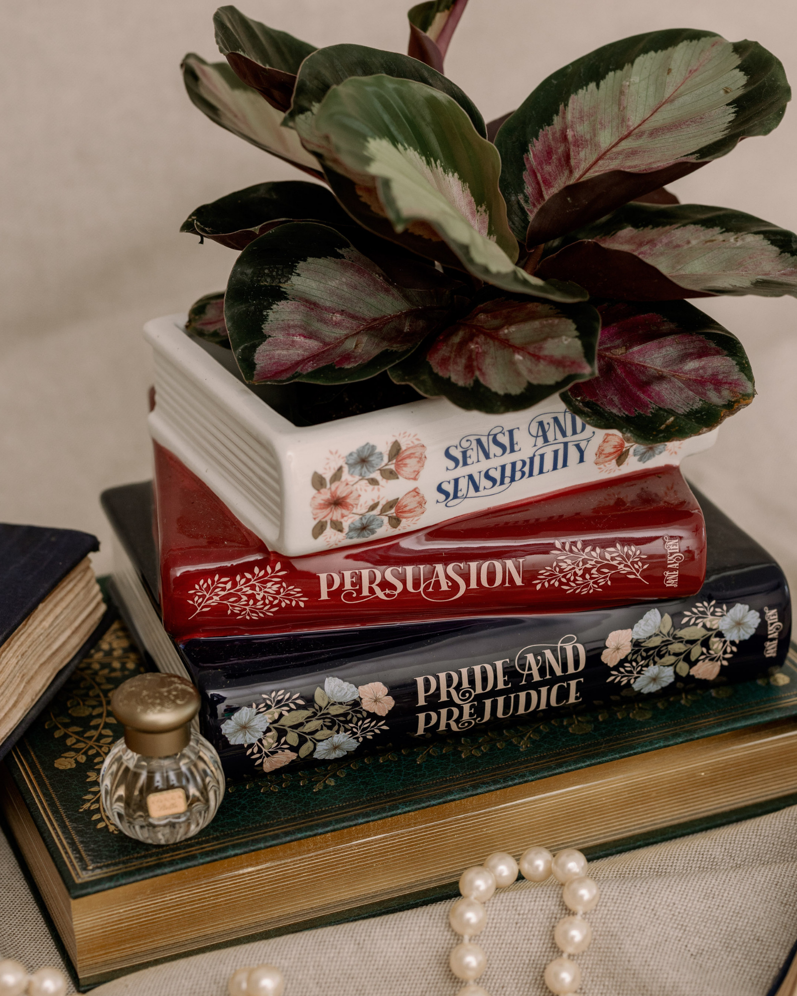 Stack of books with decorative covers, a plant, and a small bottle on a textured surface.