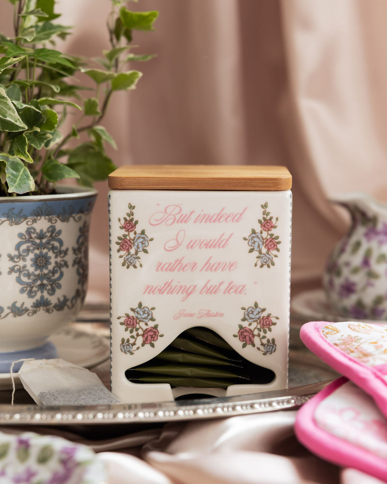 Tea bag container with floral design and text on a table with a plant and pink item in the background