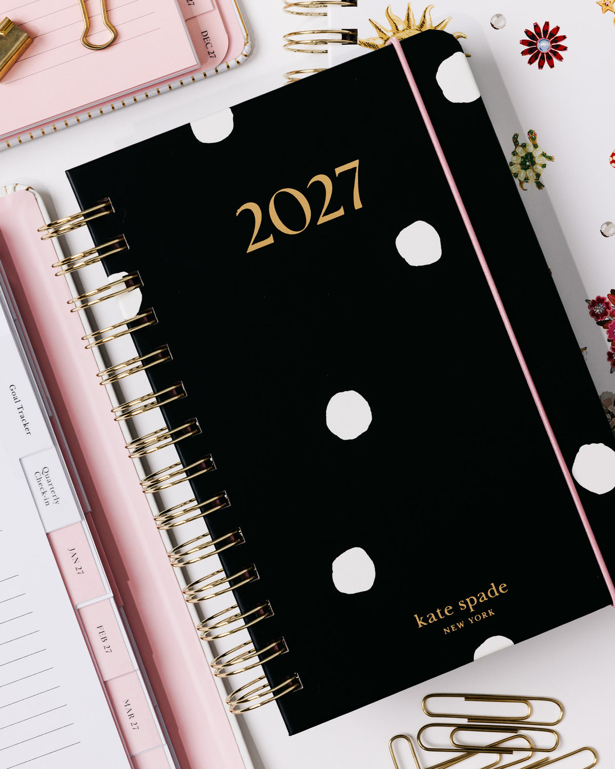 A black Kate Spade 2027 spiral planner with white polka dots and gold lettering, styled in a flat lay with pink accessories, paper clips, and decorative stickers on a white background.