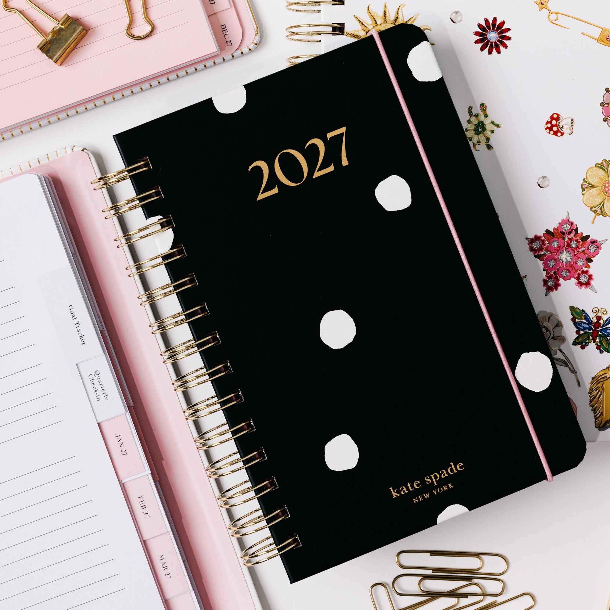 A black Kate Spade 2027 spiral planner with white polka dots and gold lettering, styled in a flat lay with pink accessories, paper clips, and decorative stickers on a white background.