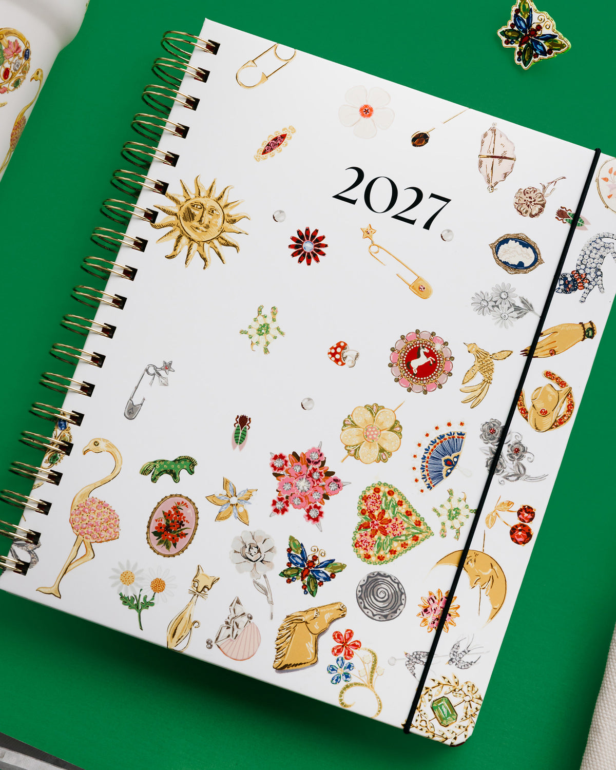 A 2027 spiral-bound planner with a white cover decorated with colorful vintage-style fashion illustrations, shown on a bright green background with styled props.