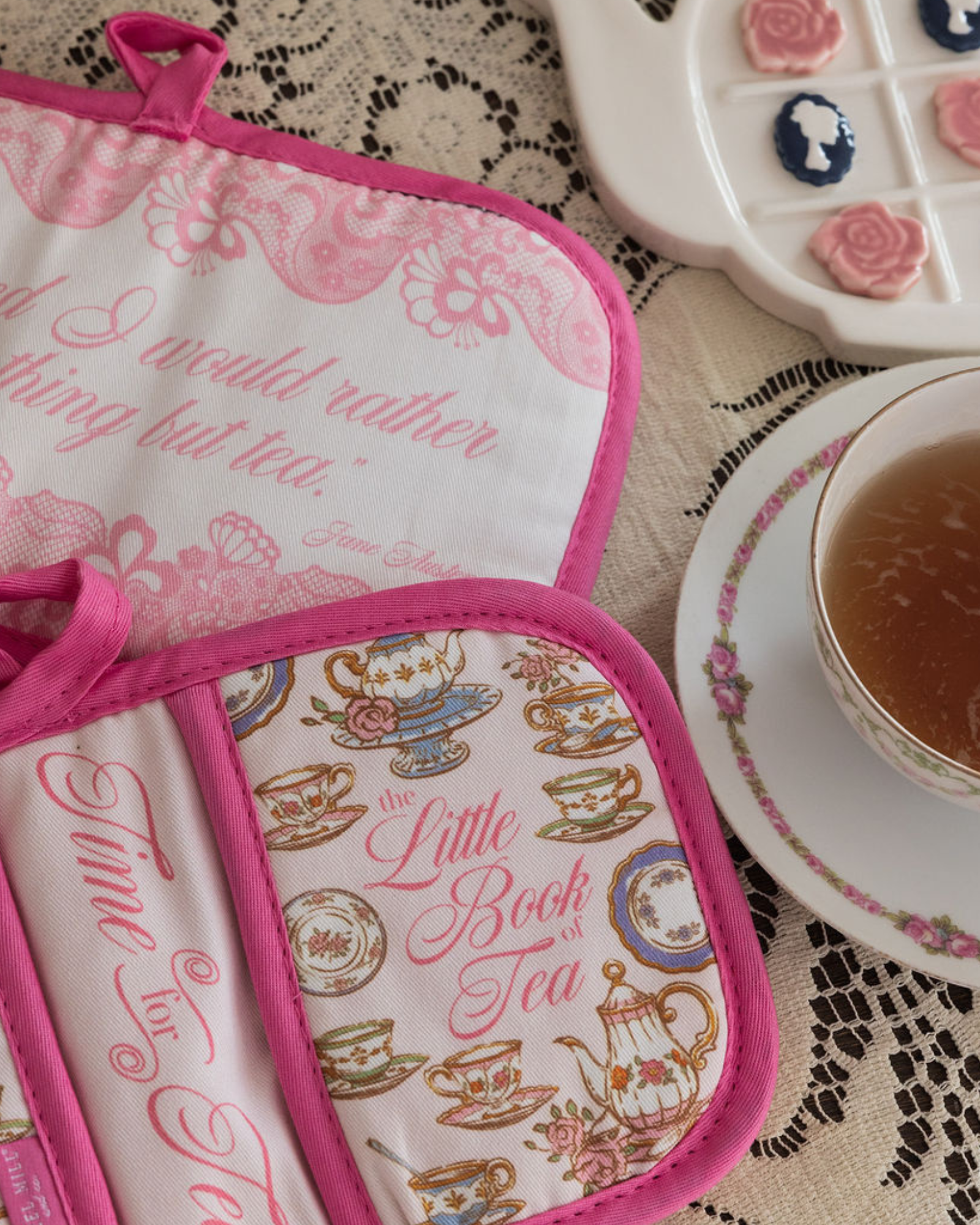 Tea-themed pot holders with decorative text and designs on a lace tablecloth with a cup of tea.