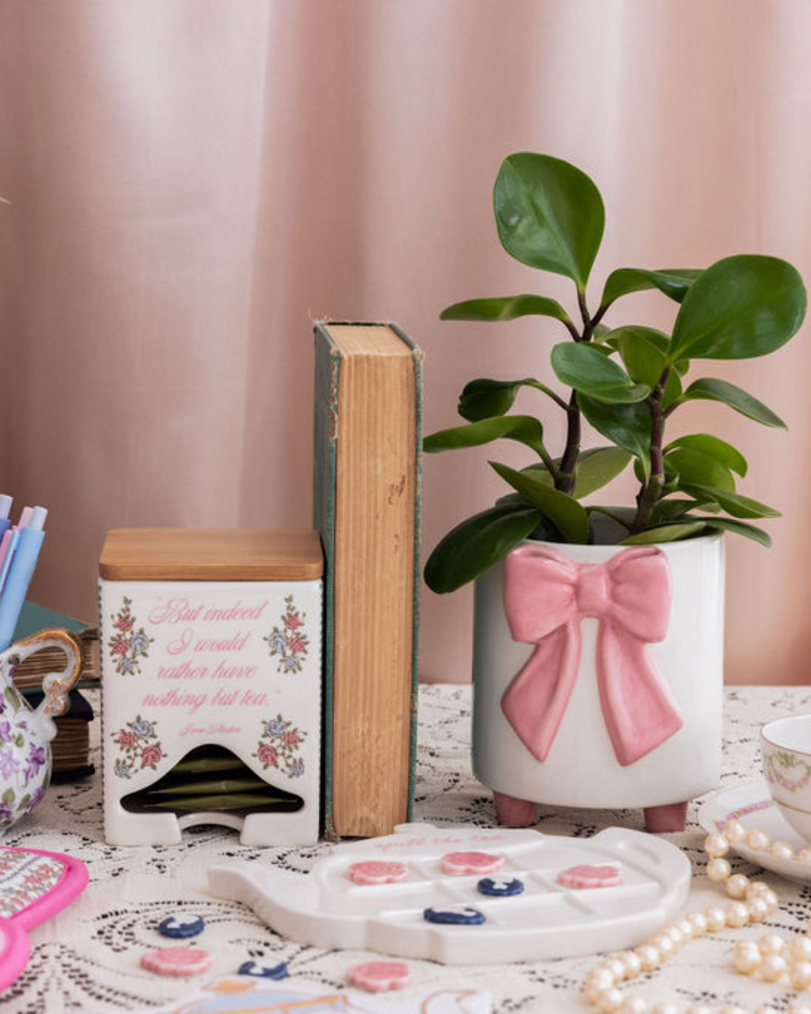 Decorative items including a potted plant with a pink bow, books, and a teapot on a table with a pink curtain background.