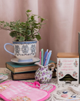 Decorative items including a teacup with plant, books, and a pink hot pad on a table.
