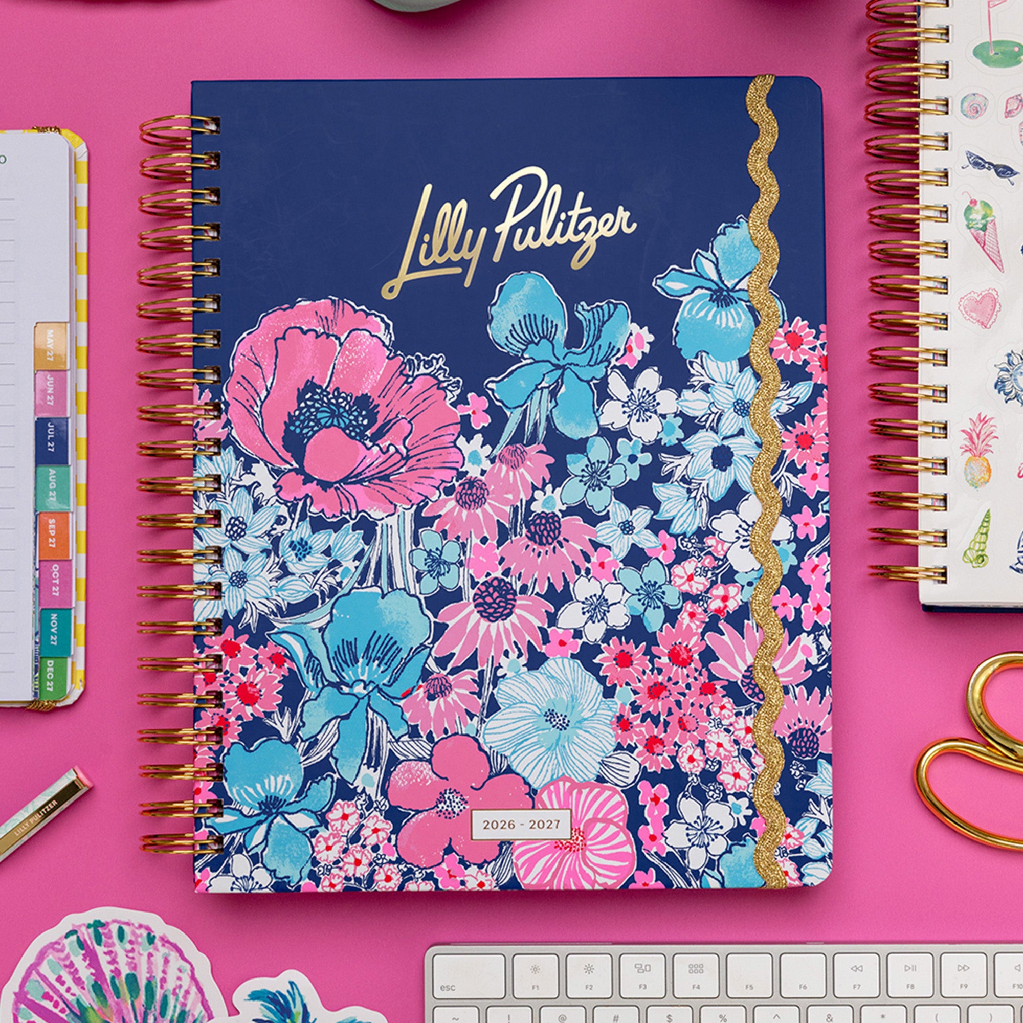 A Lilly Pulitzer 2026 daily planner with a navy blue cover, featuring a vibrant pink and blue floral pattern and gold foil lettering, styled on a bright pink desk with a keyboard, sticky notes, and other stationery.
