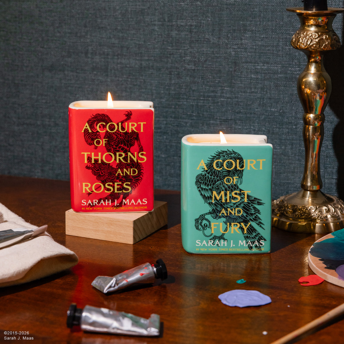 Two square candles styled like book covers, one red with gold text and black illustration of thorns and roses, the other teal with gold text and black illustration of a raven and roses. Both candles are lit and displayed on a wooden table with art supplies and a brass candlestick in the background.