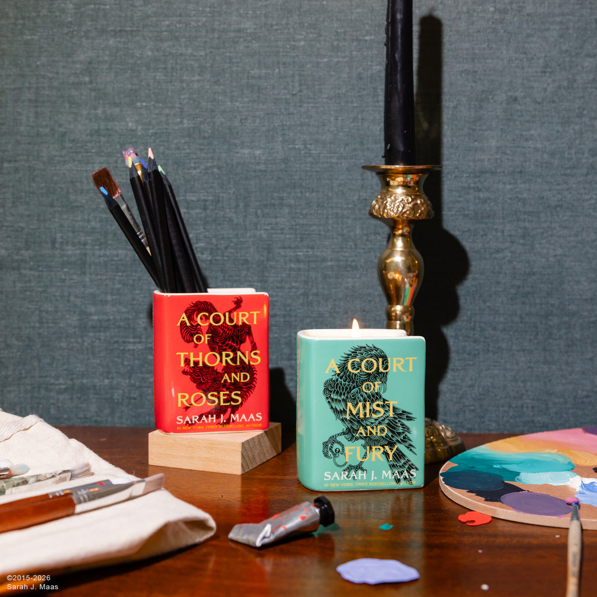Two decorative candle holders shaped like book covers, one red with a black rose and the other mint green with a black bird, displayed on a wooden table with art supplies against a textured gray wall.