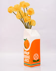 Orange juice carton vase with yellow flowers on a light gray background