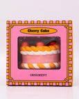 Packaged ornament cake with pink filling, orange and white striped sides, and red cherries on a white background