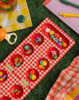 Red checkered mancala board with fruit-themed items on a grass