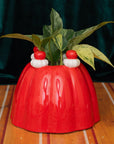 Red jelly vase planter with small red and white figures on top, containing a green plant, on a wooden surface.