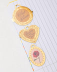 Three gold bookmarks with engraved text and a rose design on a lined paper background.