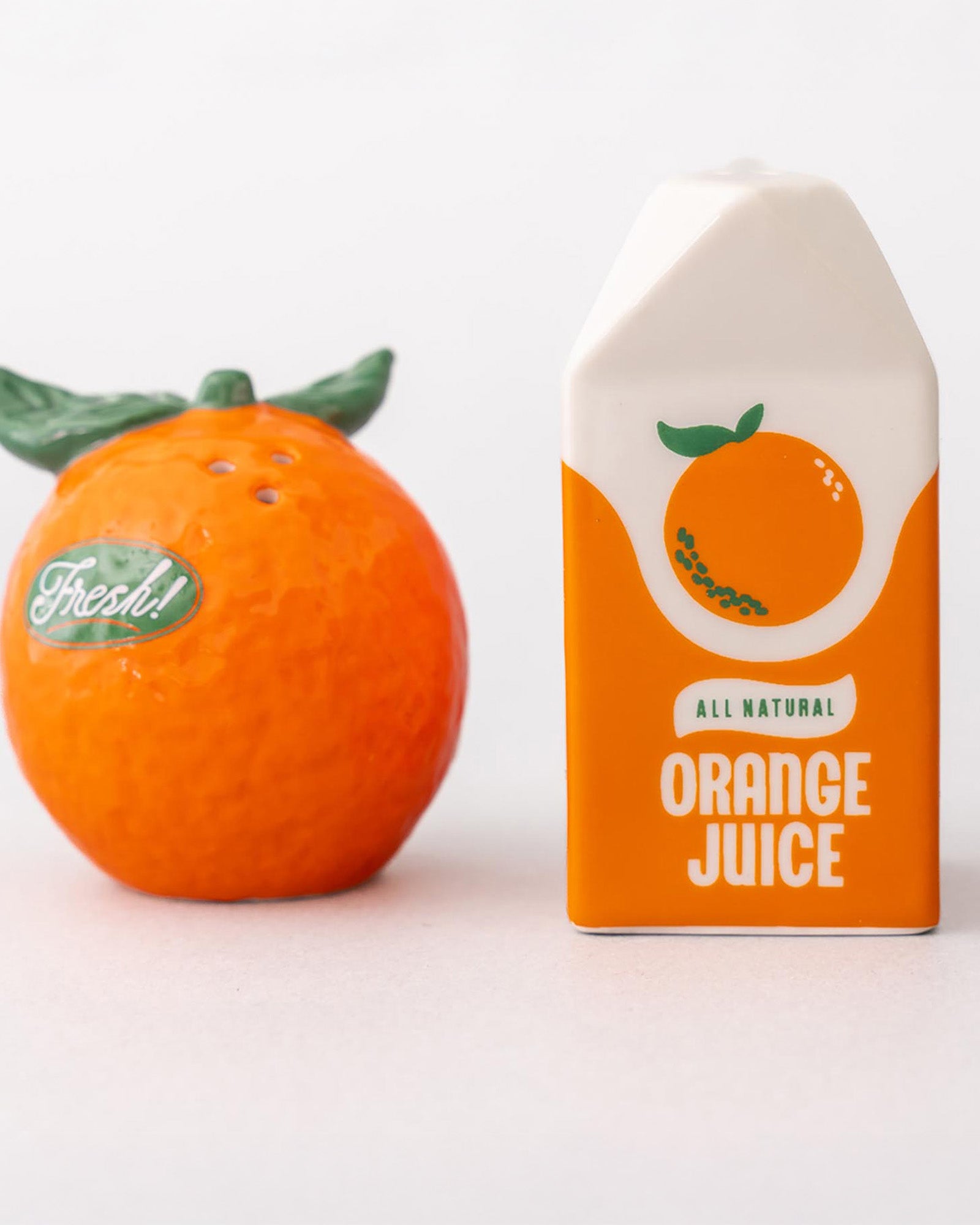 Decorative salt and pepper shaker shaped like oranges next to a box of all natural orange juice on a white background