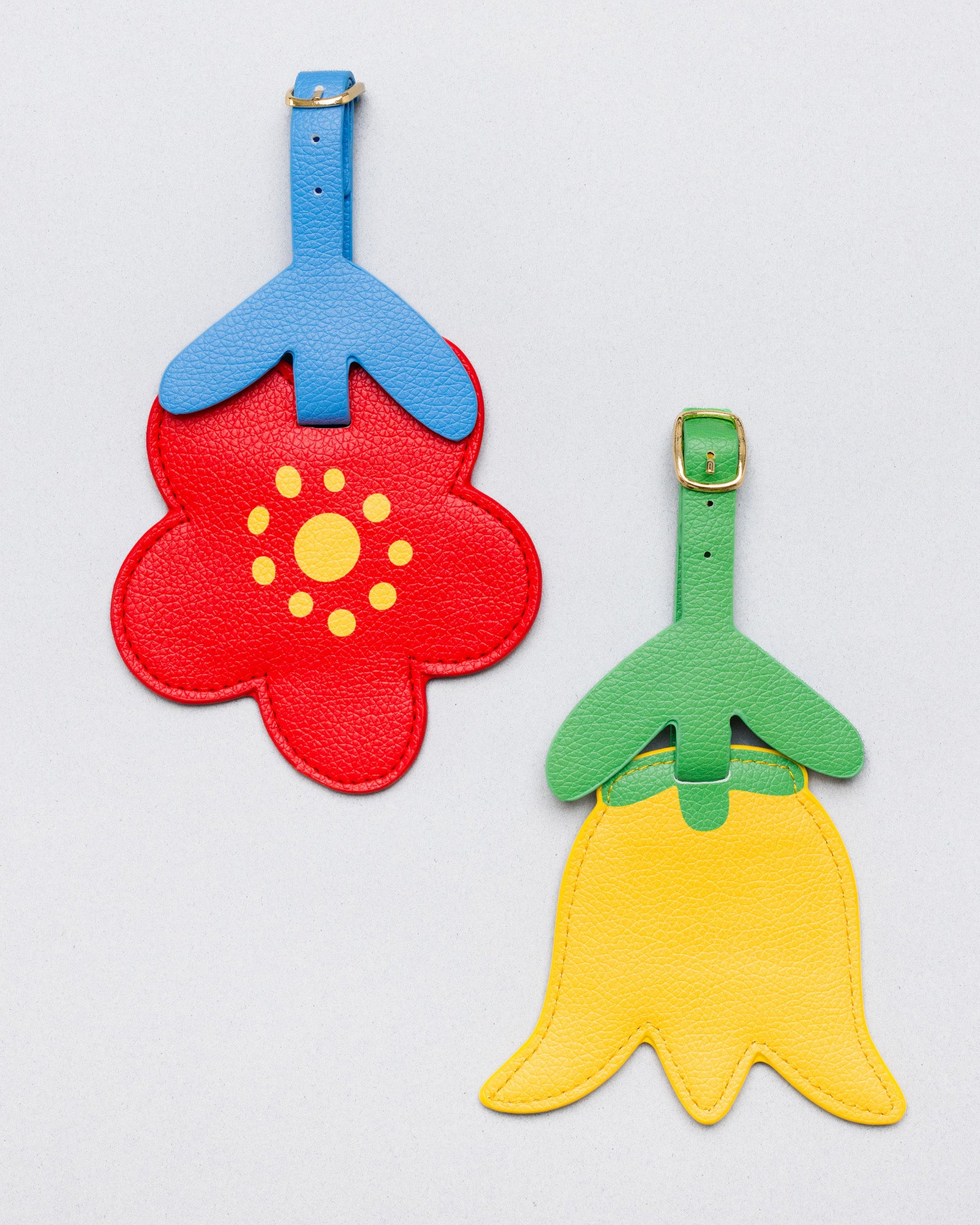 Two flower-shaped luggage tags on a light gray background. One tag is red with a blue top and yellow center, and the other is yellow with a green top and yellow center.