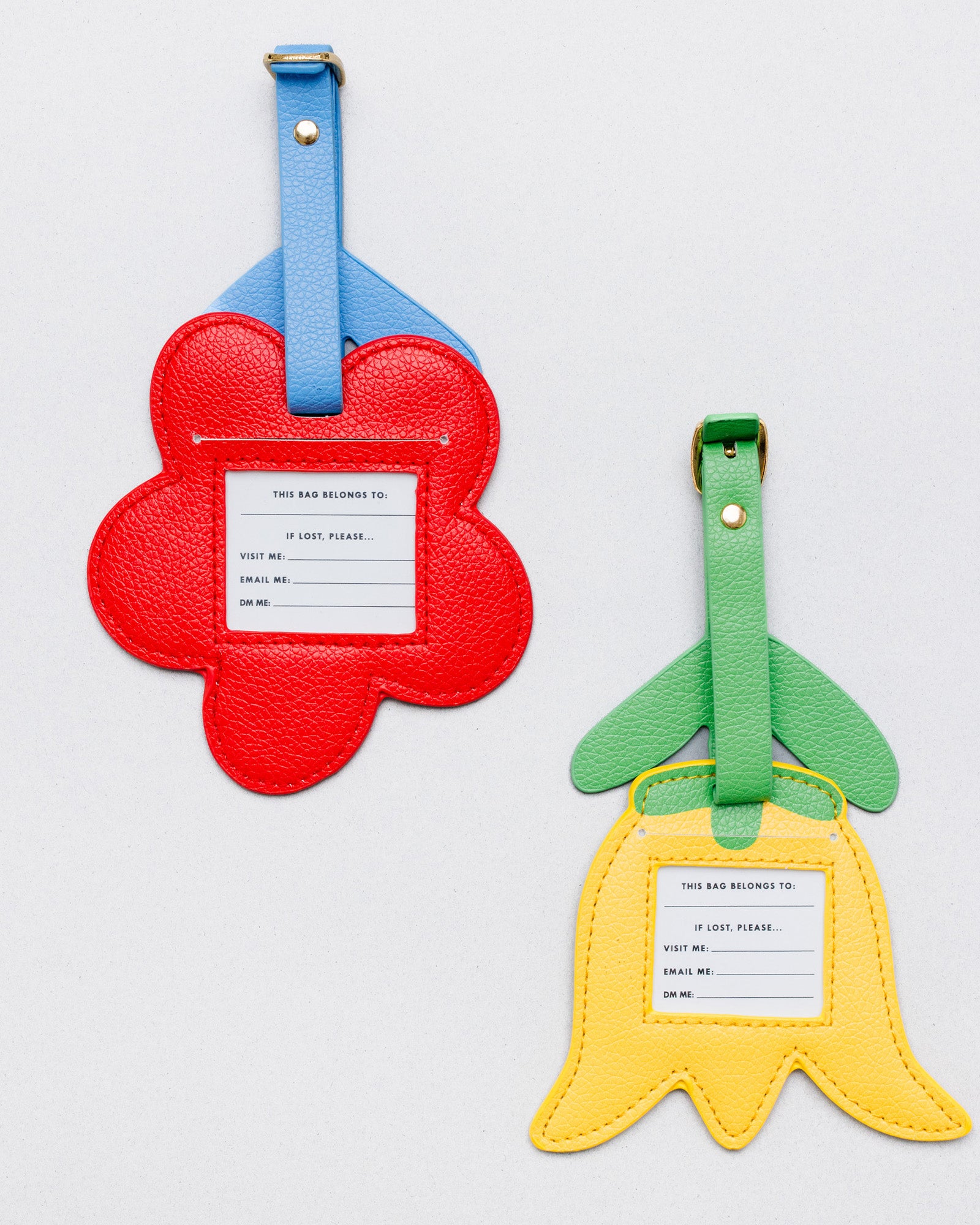 Two flower-shaped luggage tags on a light gray background. One tag is red with a blue strap and a white insert, and the other is yellow with a green strap and a white insert.