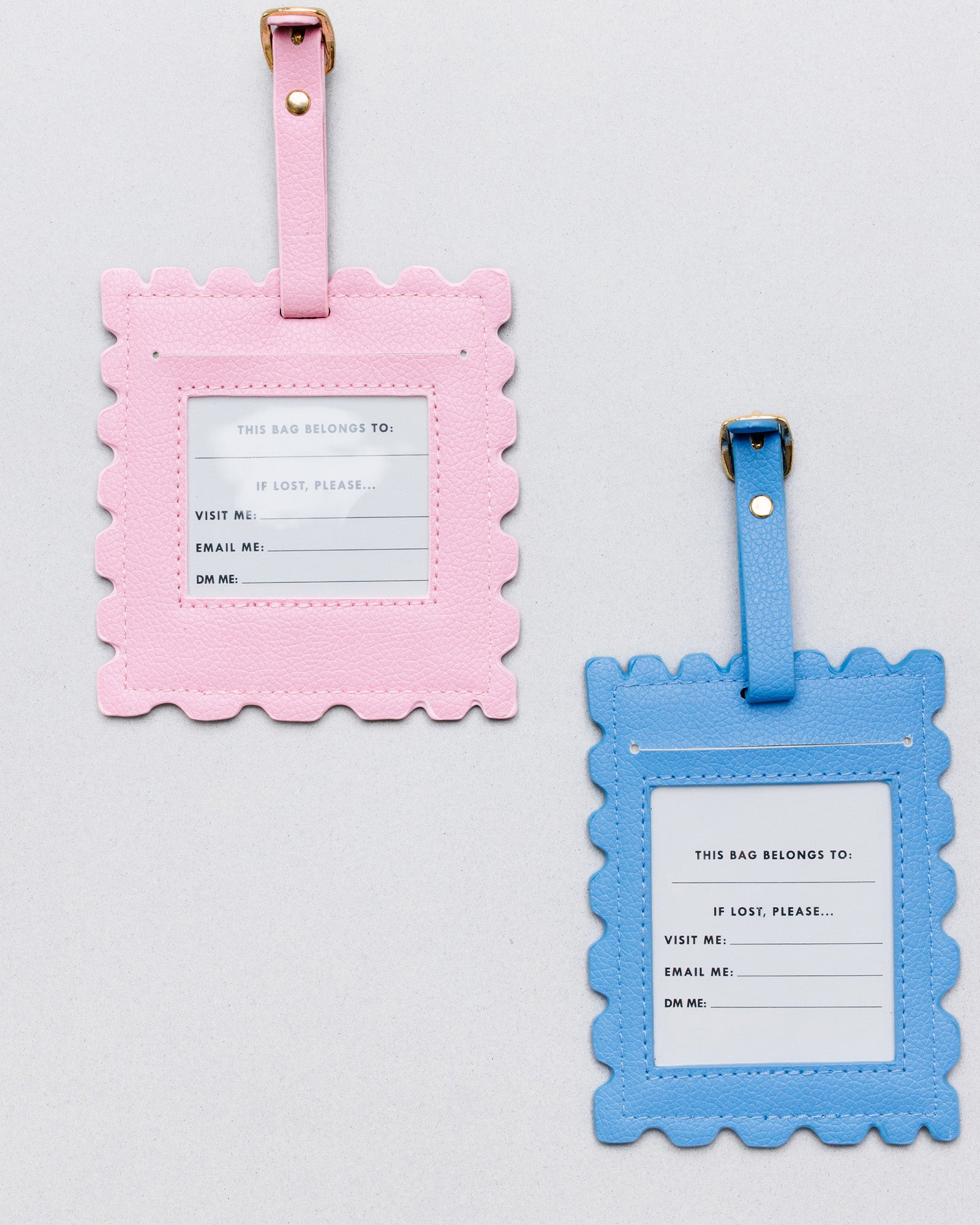 Two scalloped luggage tags on a light gray background, one in pink and one in blue, each with a white insert card and gold-tone metal hardware.