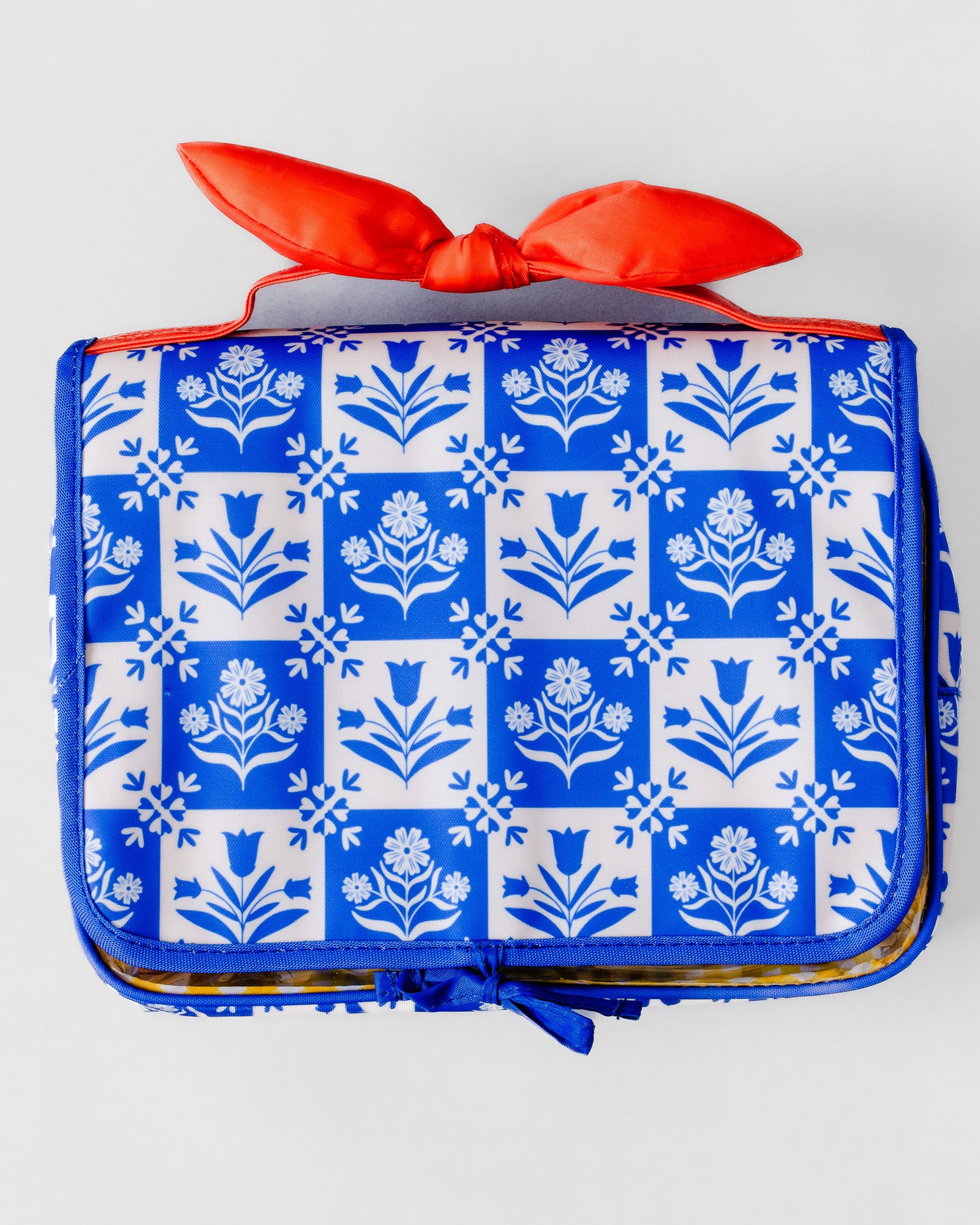 A rectangular zippered travel or toiletry bag with a blue and white floral tile pattern, featuring a bright orange-red satin ribbon tied in a bow on top, photographed against a light gray background.