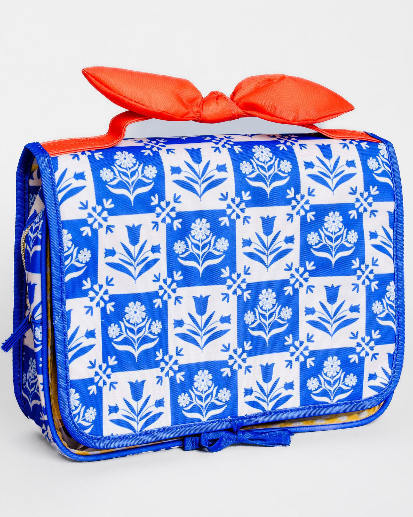 A rectangular soft-sided bag with a blue and white floral block-print pattern, featuring a bright orange-red satin bow on top and a matching blue zipper pull, shown on a light gray background.