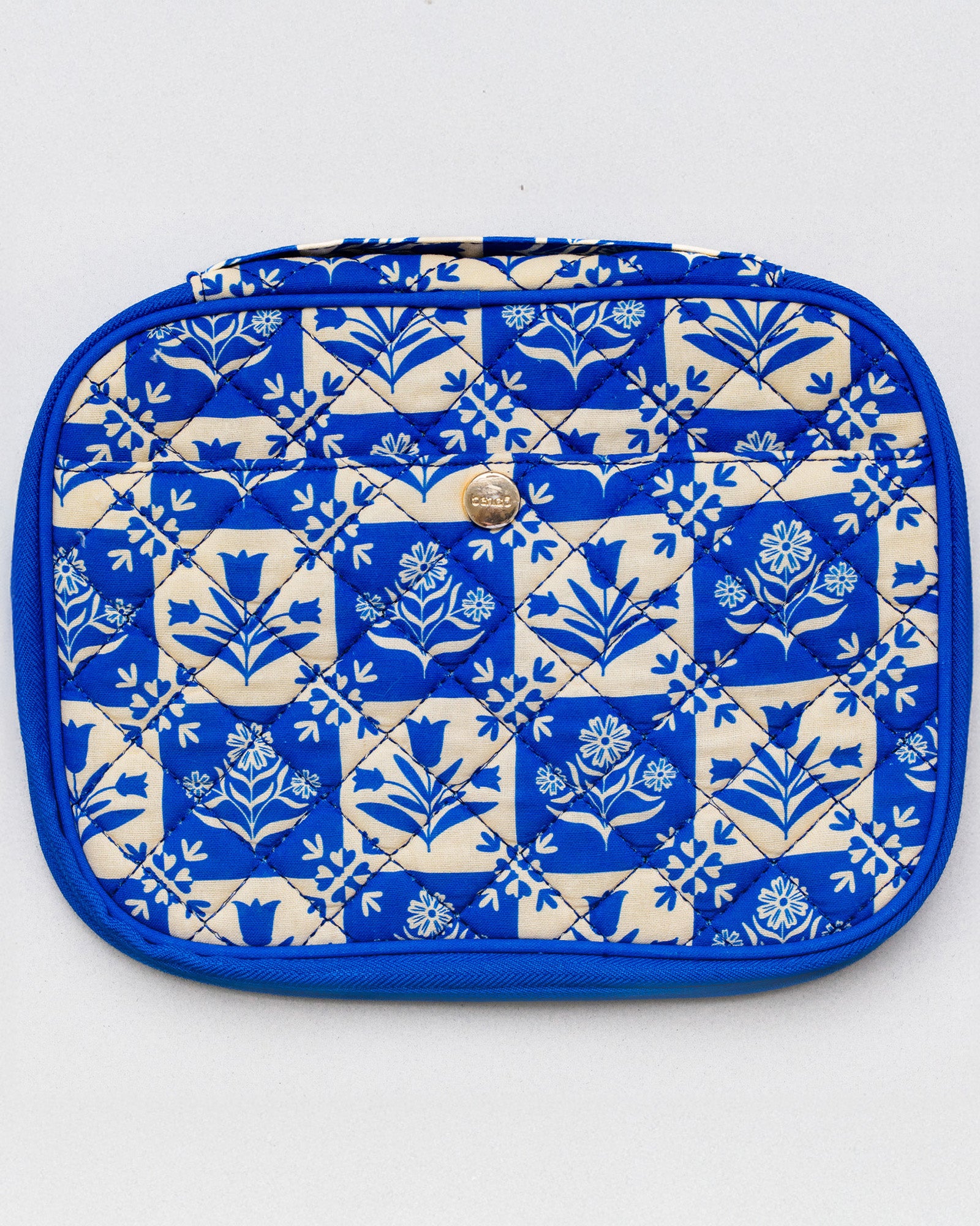 A quilted cosmetic pouch with a blue and cream floral block-print pattern, featuring a small gold metal logo on the front.