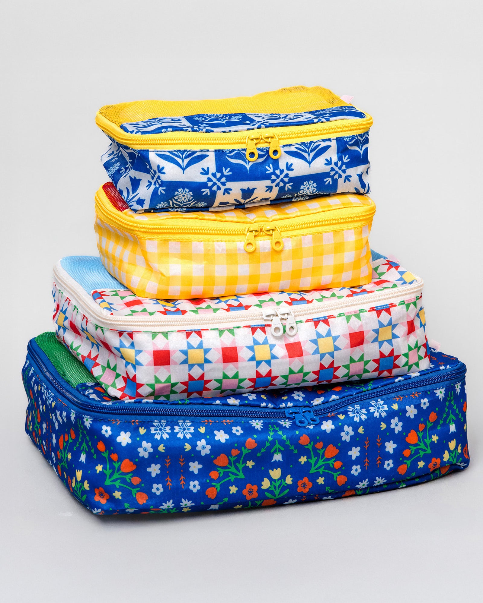 A stack of four colorful zippered fabric storage bags in various sizes, featuring different patterns including blue floral, yellow gingham, and multicolor geometric, set against a light gray background.