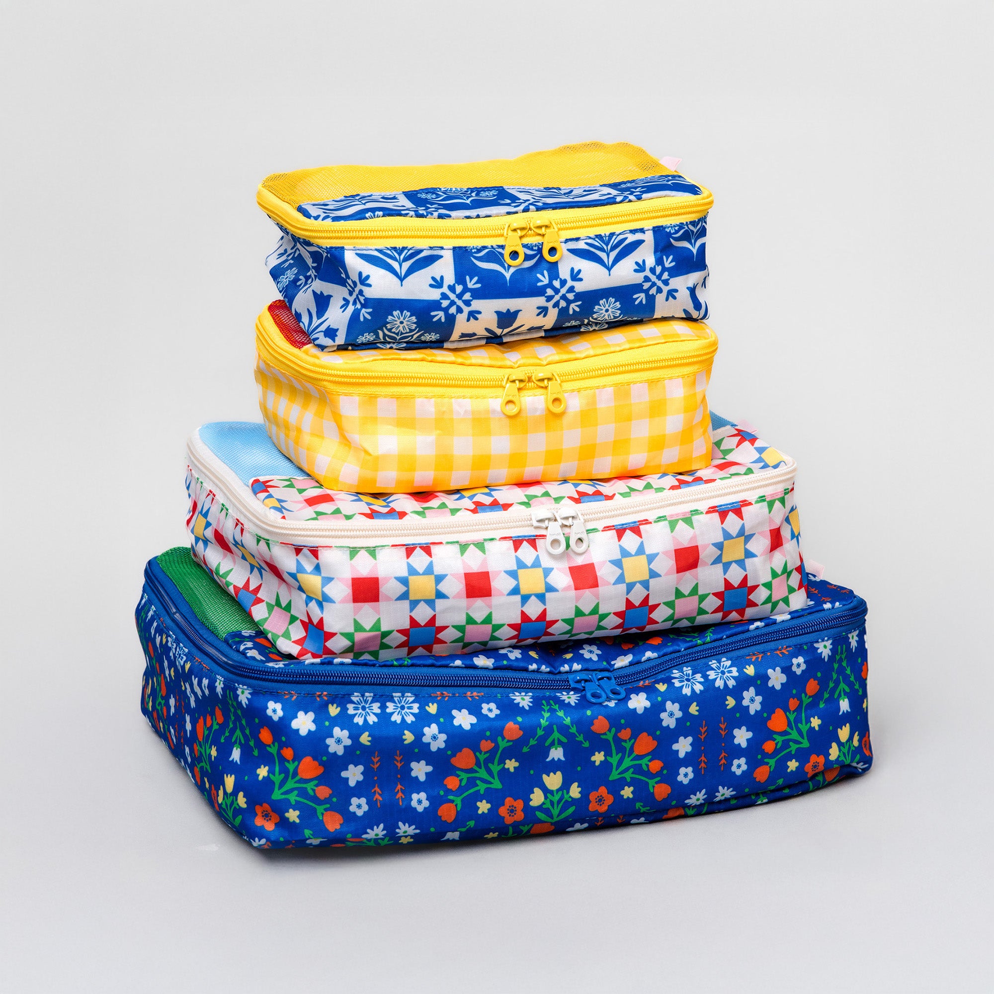 A stack of four colorful zippered fabric storage bags in various sizes, featuring different patterns including blue floral, yellow gingham, and multicolor geometric, set against a light gray background.