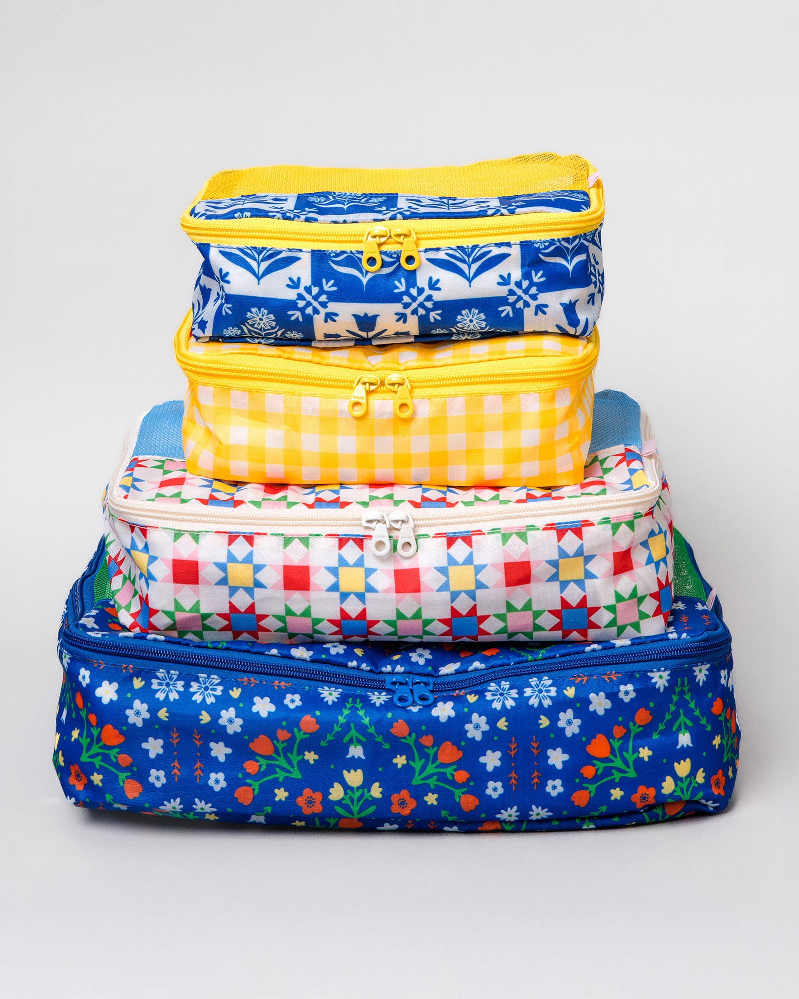 A stack of four colorful zippered fabric storage bags with various patterns including gingham, floral, and geometric, set against a plain light gray background.
