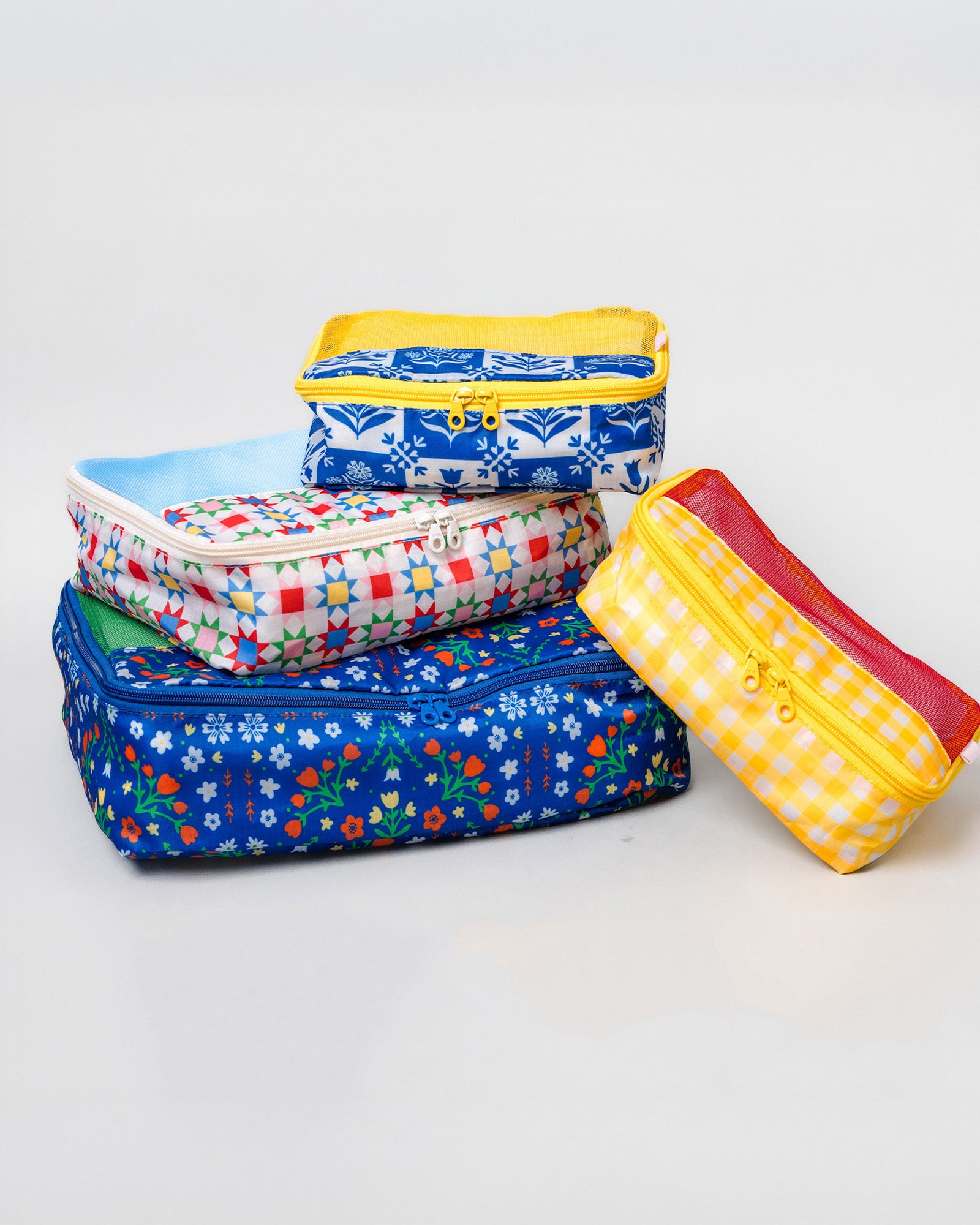 Four fabric zippered pouches stacked and leaning on a light gray studio background. The set includes blue floral, red-and-white gingham, yellow-and-white gingham, and blue-and-white patterned pouches with yellow and red zippers.