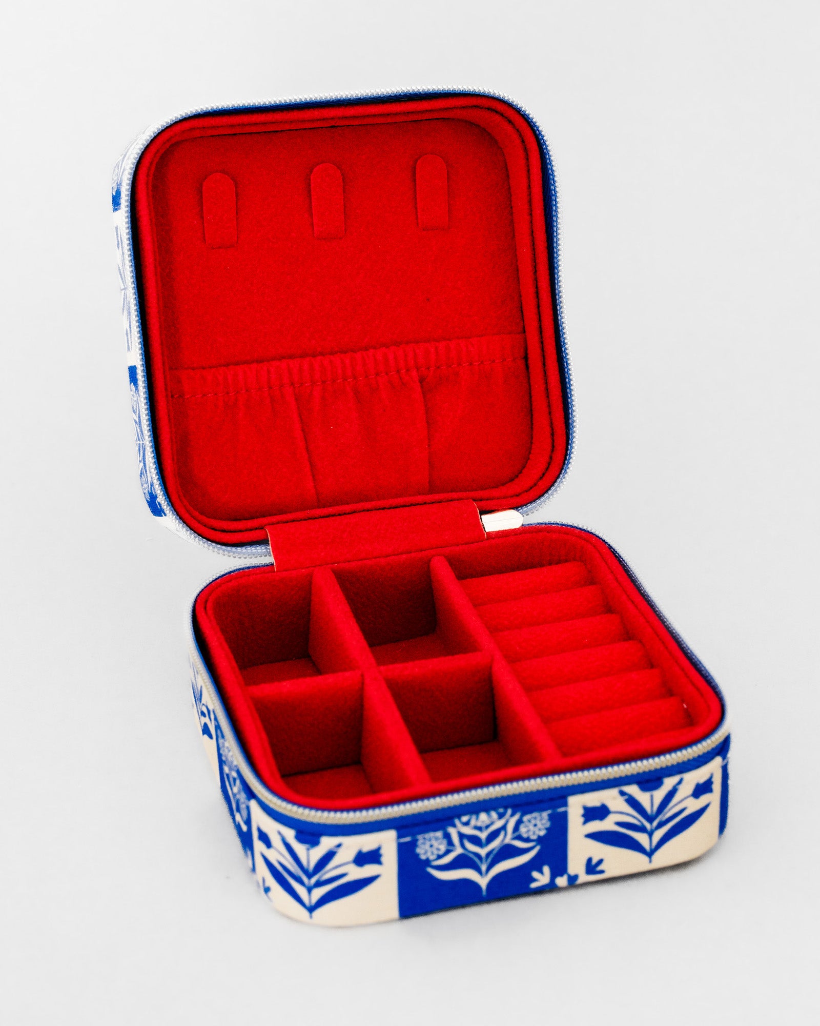 An open fabric jewelry box with a red velvet interior and a blue and white floral patterned exterior, shown on a light gray background.