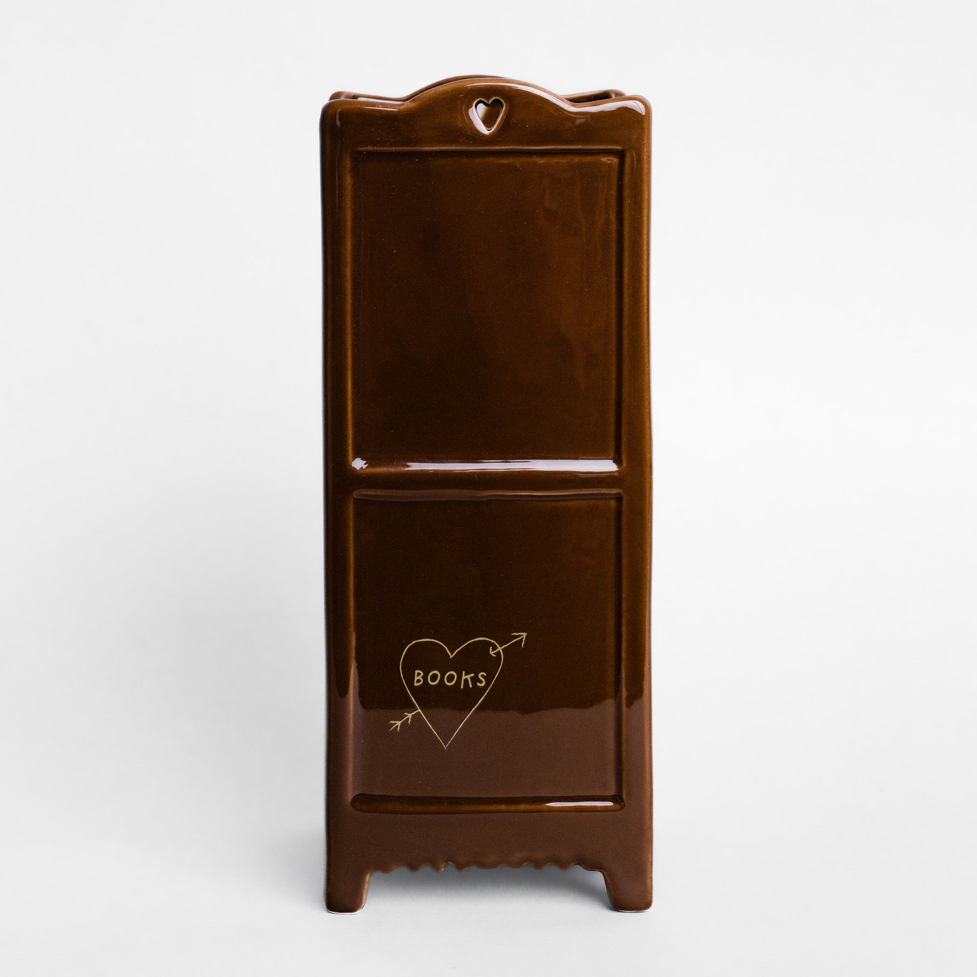 A glossy dark brown ceramic plant pot shaped like a miniature bookshelf featuring a small heart-shaped cutout at the top and a gold heart-and-arrow graphic with the word 'BOOMS' on the lower door, set against a plain light gray background.