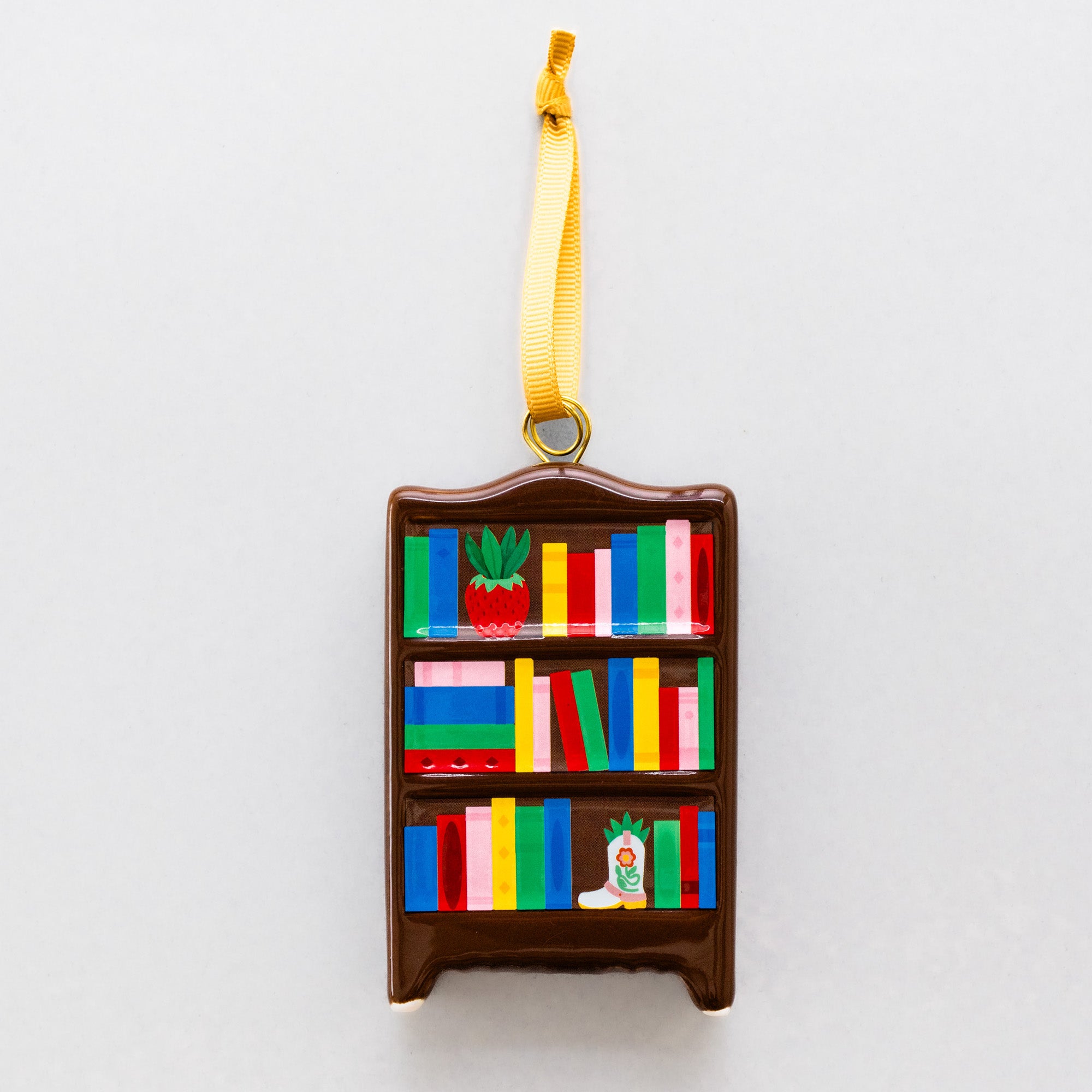 A brown bookshelf-shaped Christmas ornament with colorful book spines and a yellow hanging ribbon, shown against a plain light gray background.