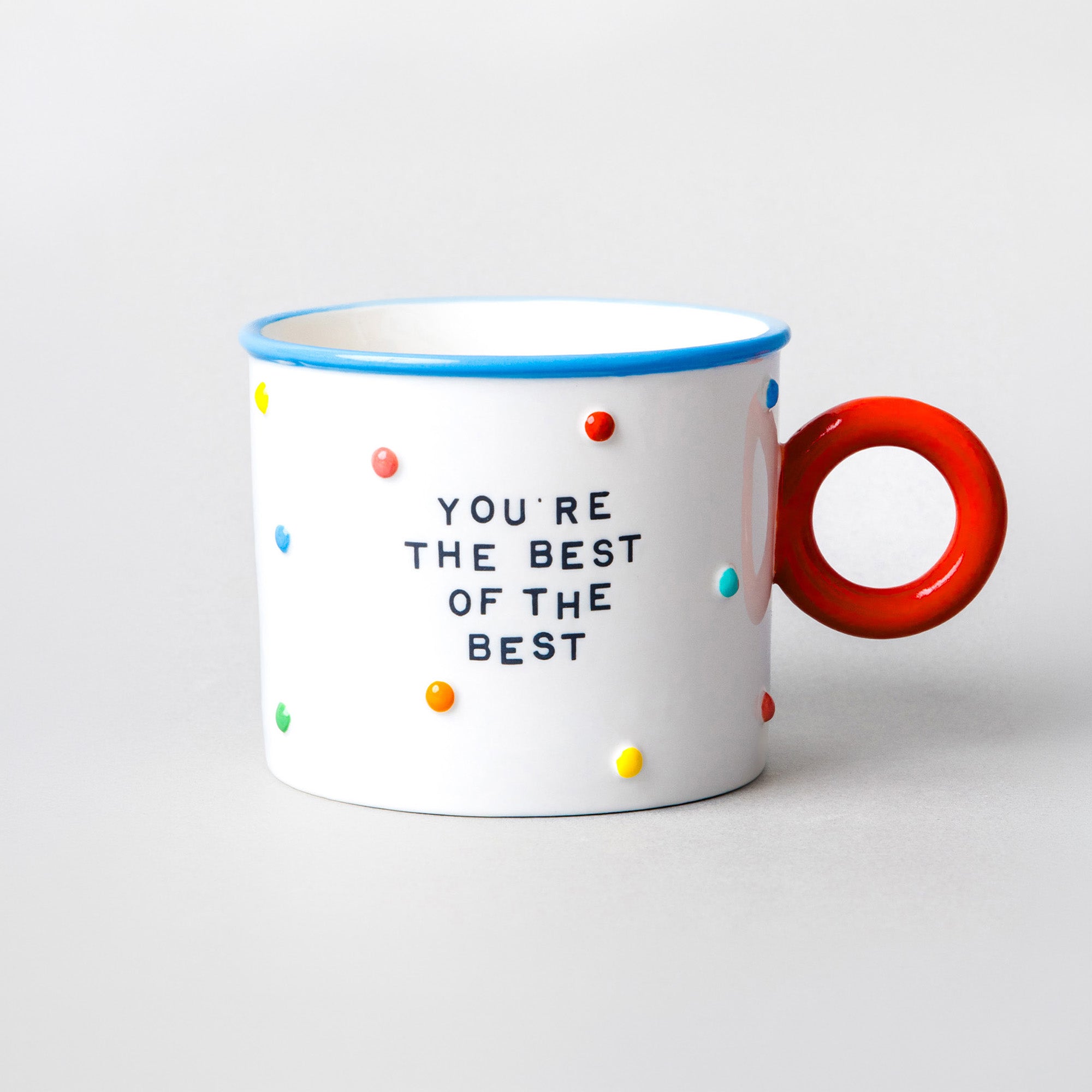 A white ceramic mug with a bright red handle, a blue rim, and multicolored polka dots. The front features the text 'YOU'RE THE BEST OF THE BEST' in black lettering.