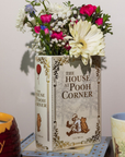 Floral arrangement in a 'The House at Pooh Corner' book on a table with a mug and jar.