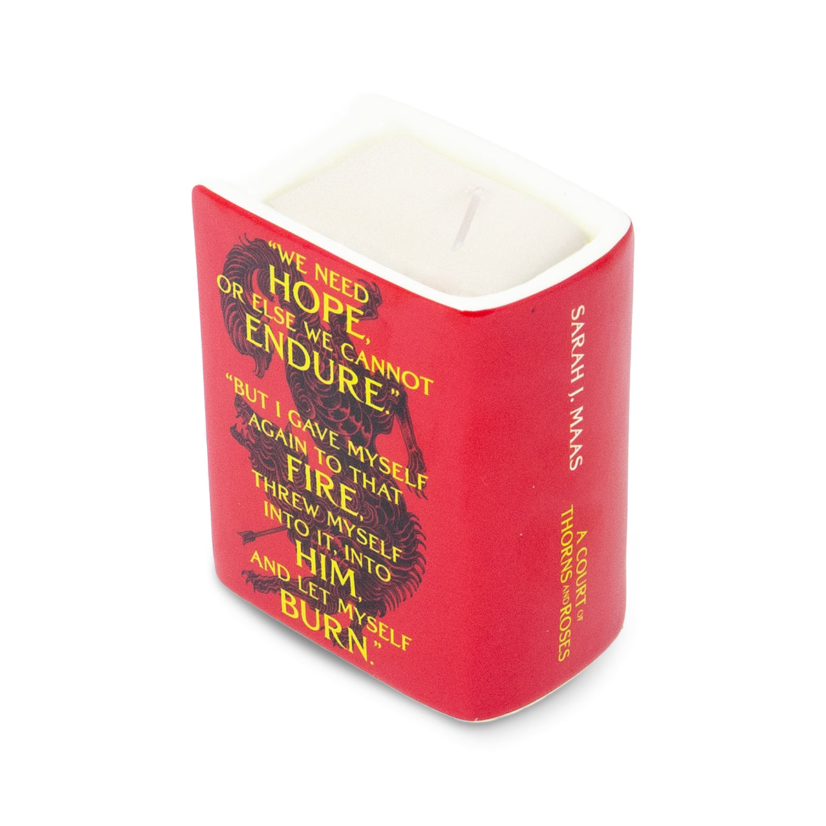 A red square candle in a white ceramic container with a white wick, featuring a black creature illustration and yellow text on the front, photographed on a white background.