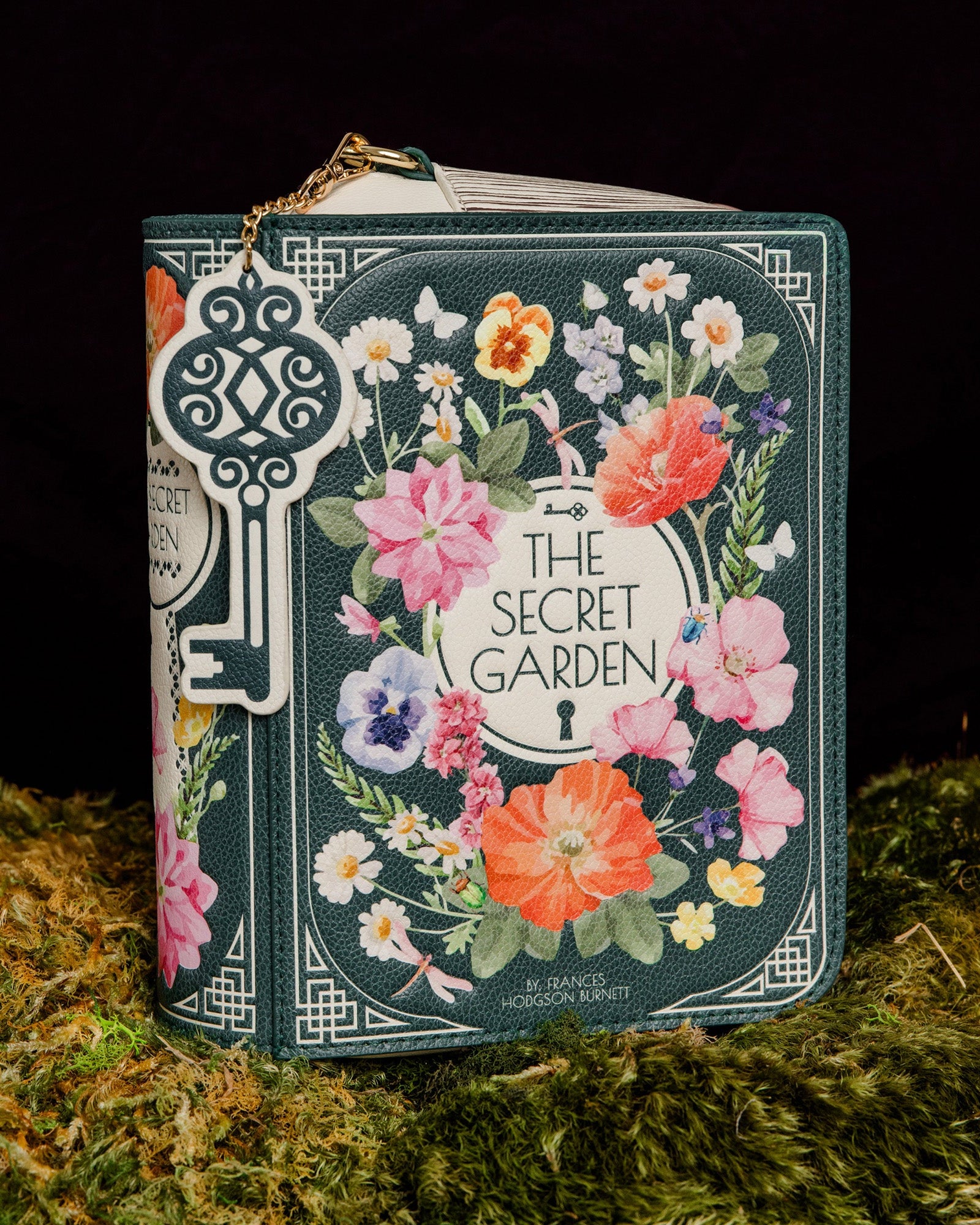 A decorative book-shaped handbag styled like 'The Secret Garden' book cover, featuring a floral wreath illustration and a large ornate key charm, displayed on moss against a black background.