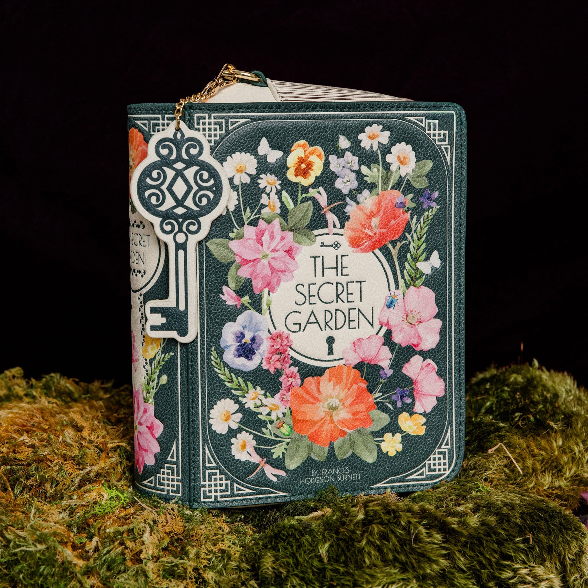 A decorative book-shaped handbag styled like 'The Secret Garden' book cover, featuring a floral wreath illustration and a large ornate key charm, displayed on moss against a black background.