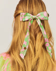 Floral hair bow in green and pink on a person with long blonde hair against a white background