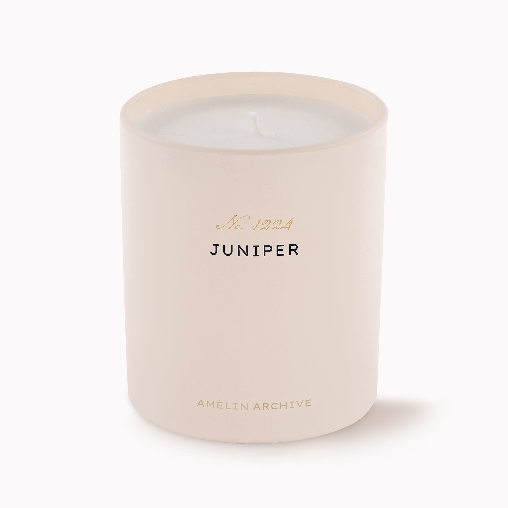 A matte blush pink glass candle jar with a white wax fill and a single wick. The front features black text reading 'JUNIPER' with pink and gold foil accents above and below, on a clean white background.