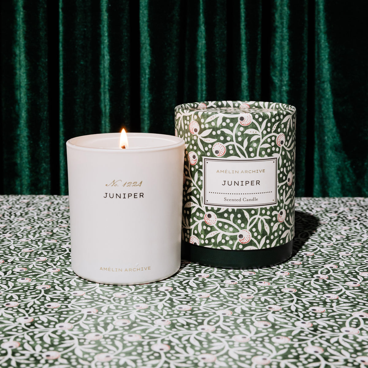 A white glass candle jar labeled 'Amelin Archive Juniper' with a small flame, standing beside its patterned green and white cylindrical box on a matching patterned surface in front of a dark green velvet curtain.