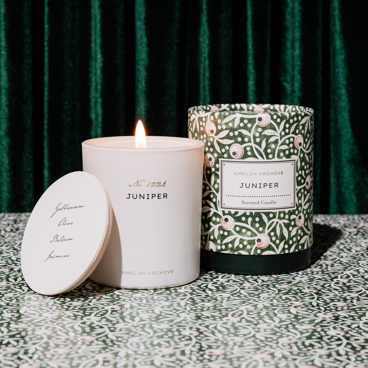 lit Amëlin Archive Juniper scented candle in a cylindrical glass container with a green and white botanical pattern packaging and a white label in the center.