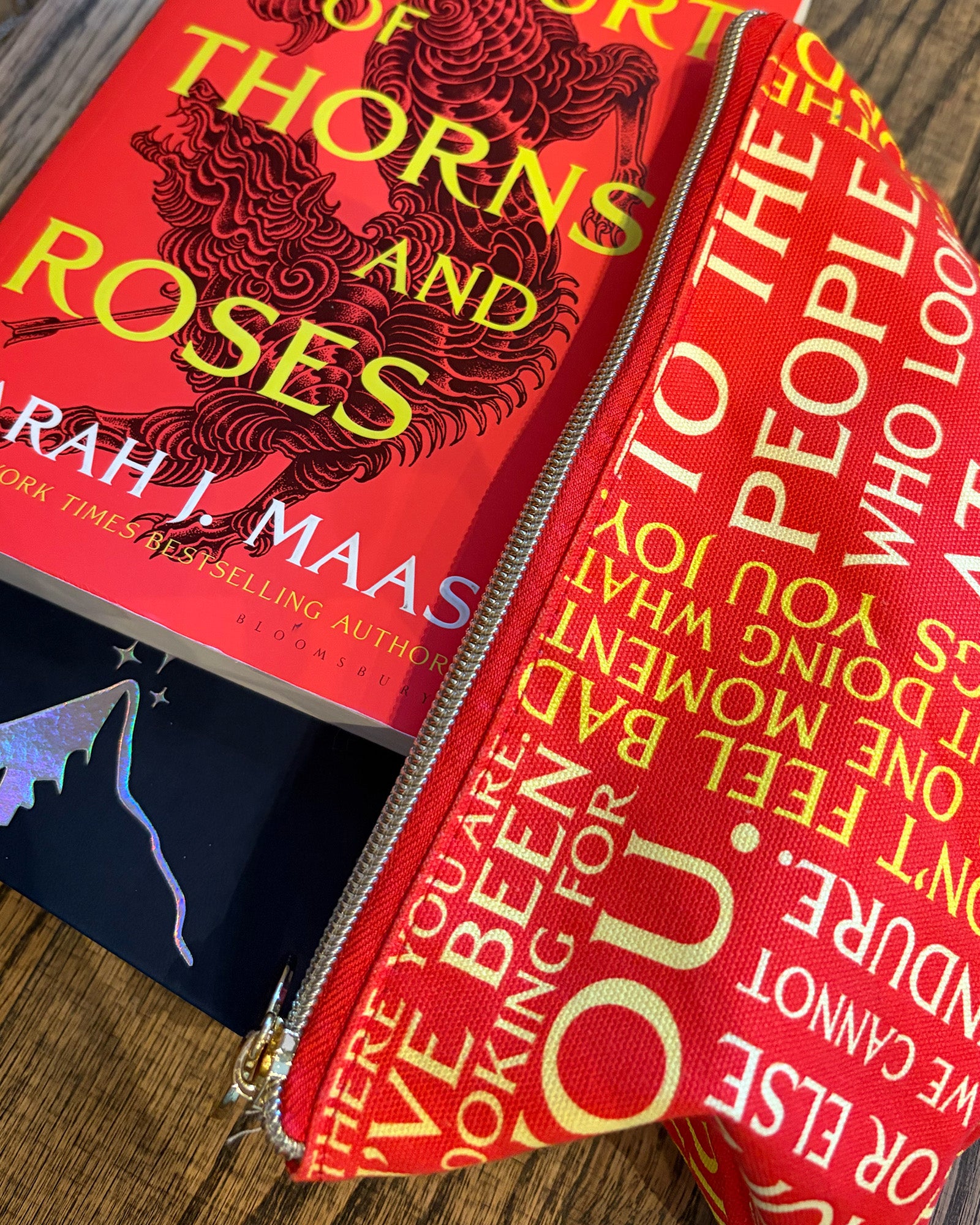 A red zippered fabric pouch printed with white and yellow text from the book 'Thorns and Roses' by Sarah J. Maas, shown open on a wooden surface next to the book.