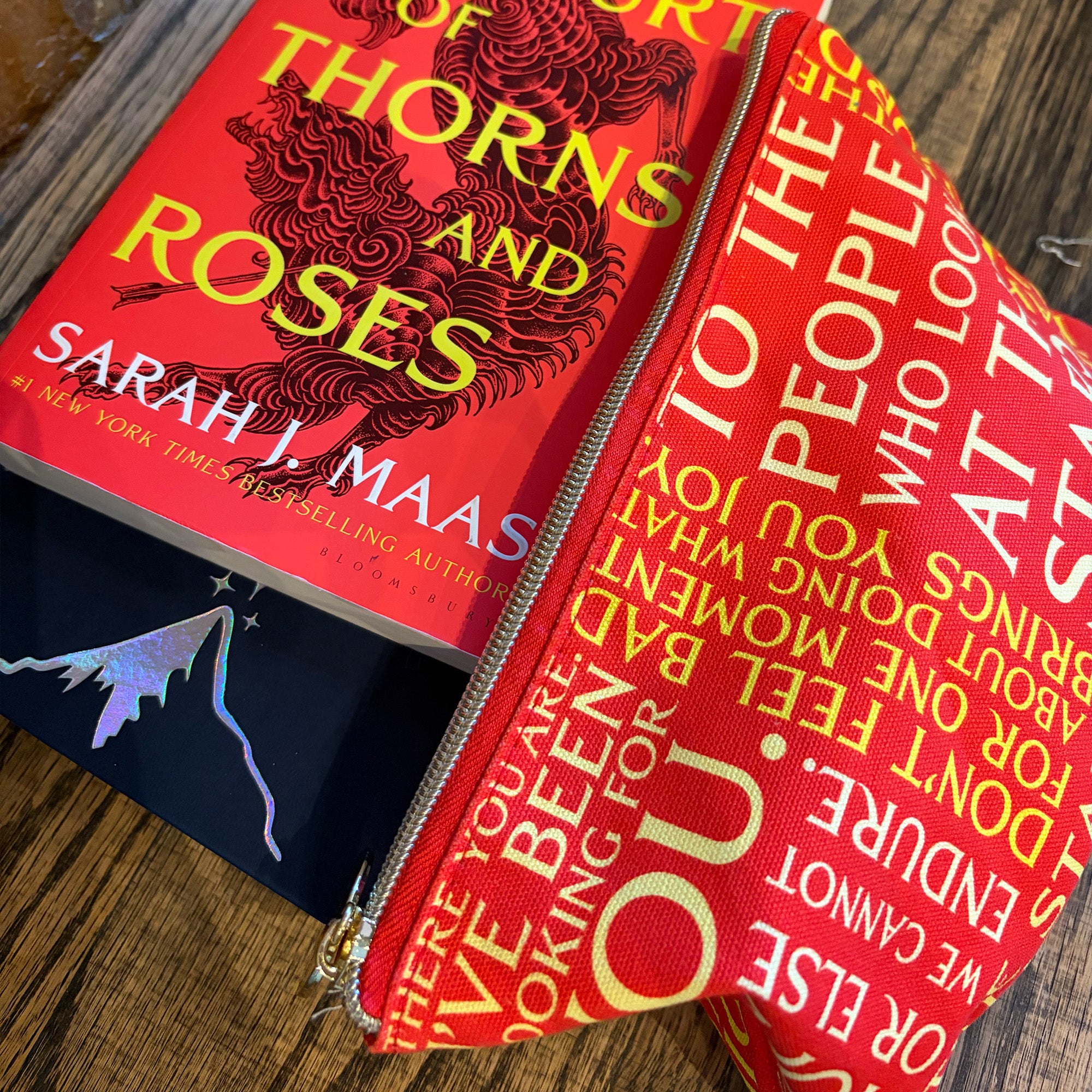 A red zippered fabric pouch printed with white and yellow text from the book 'Thorns and Roses' by Sarah J. Maas, shown open on a wooden surface next to the book.