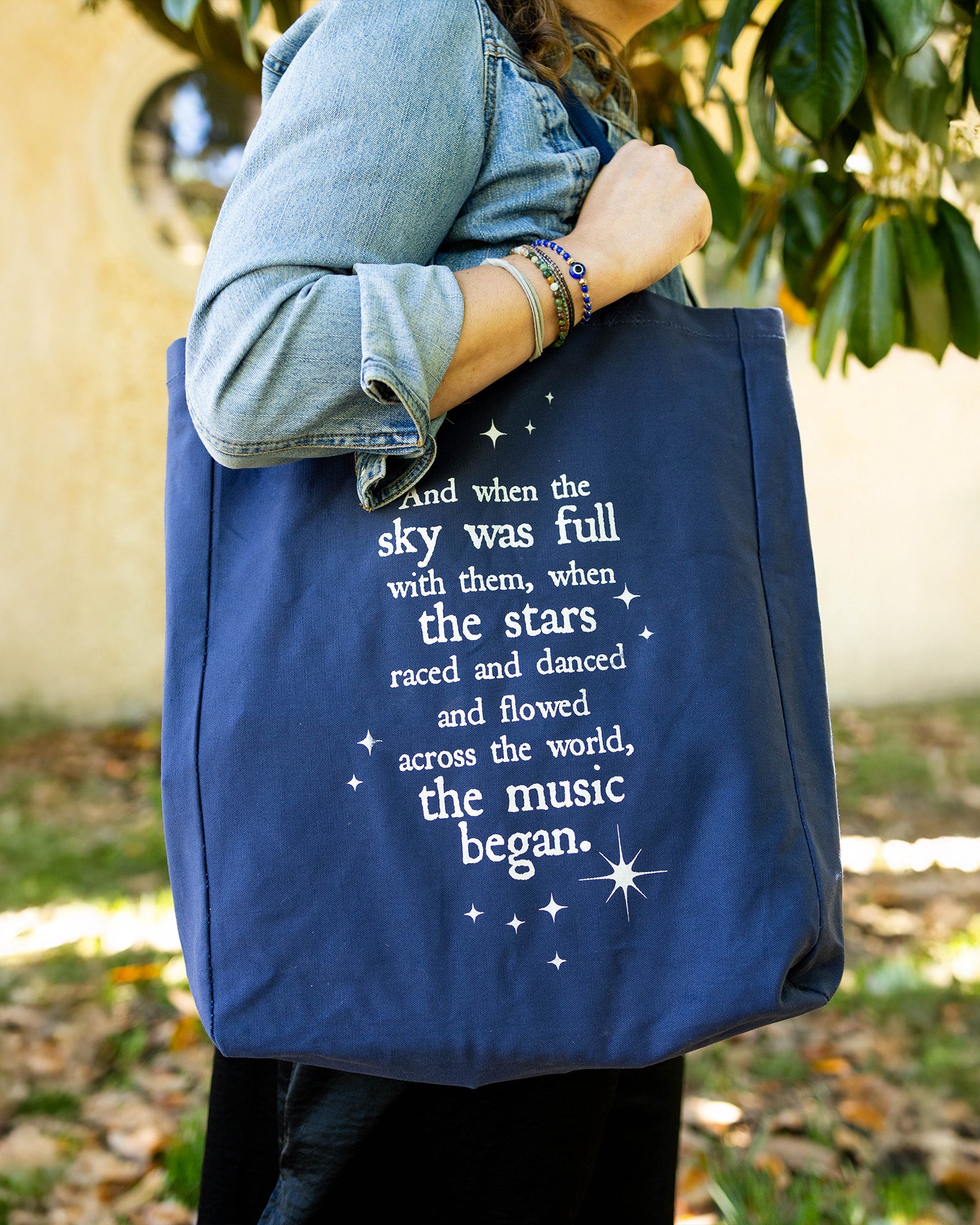 model carrying navy tote that says 'AND WHEN THE SKY WAS FULL WITH THEM, WHEN THE STARS RACES AND DANCED AND FLOWERS ACROSS THE WORLD, THE MUSIC BEGAN'