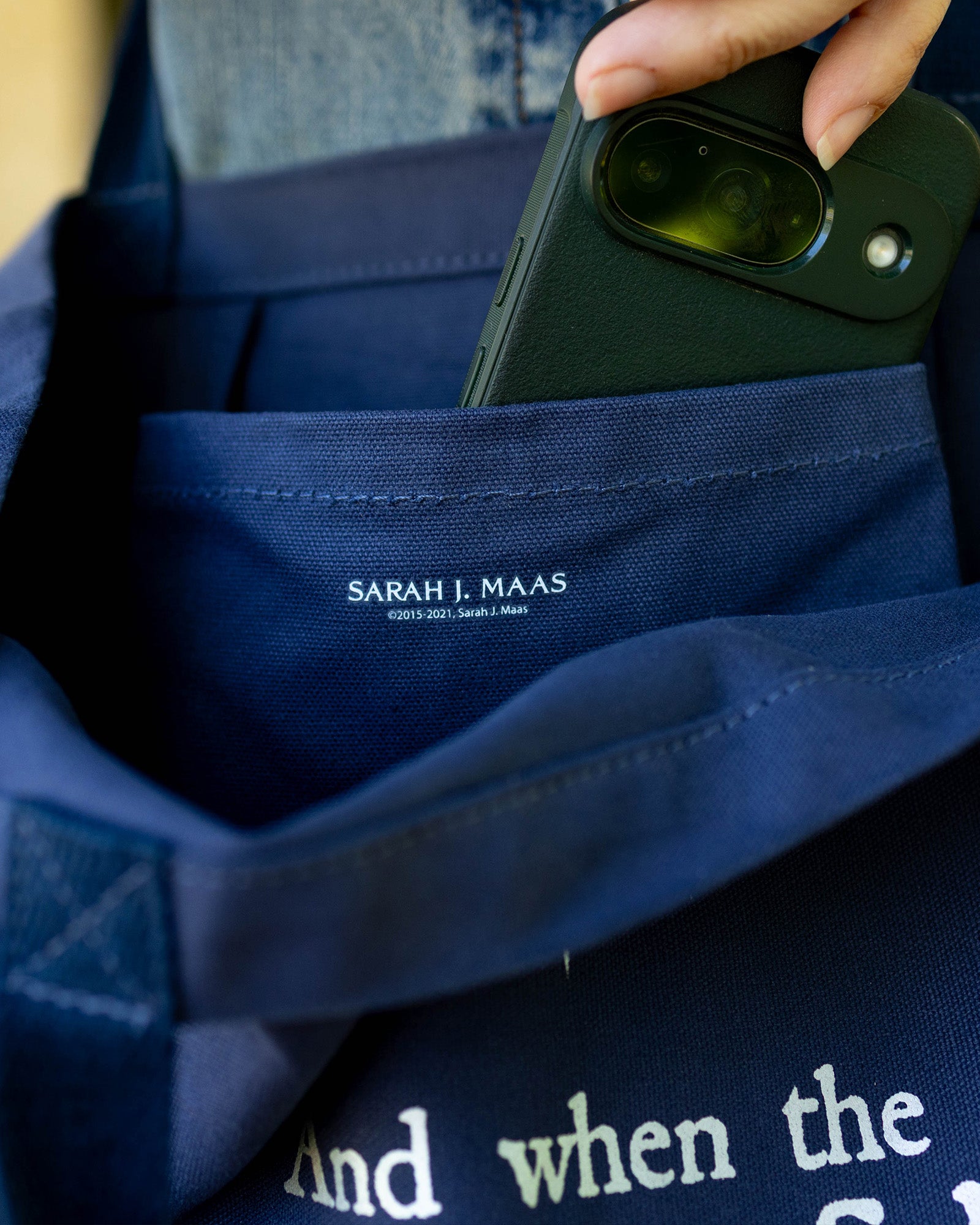 Person holding a phone over a blue tote with 'Sarah J. Maas' branding.